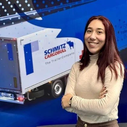 Smiling young woman with arms crossed stands in front of a Schmitz Cargobull trailer display, ready to welcome you.