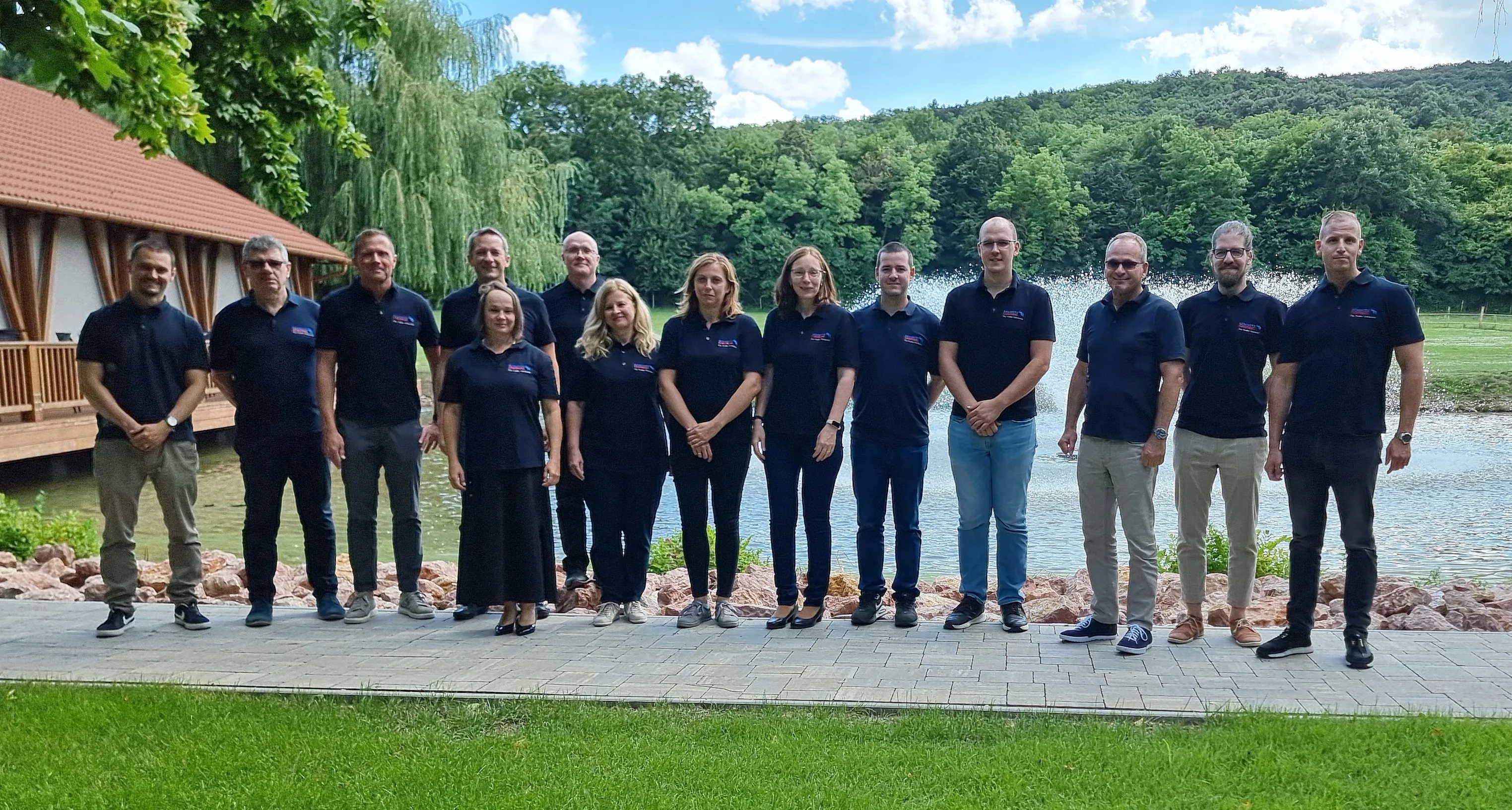 A friendly group of Schmitz Cargobull AG team members posing outdoors by a scenic pond, wearing matching branded shirts.