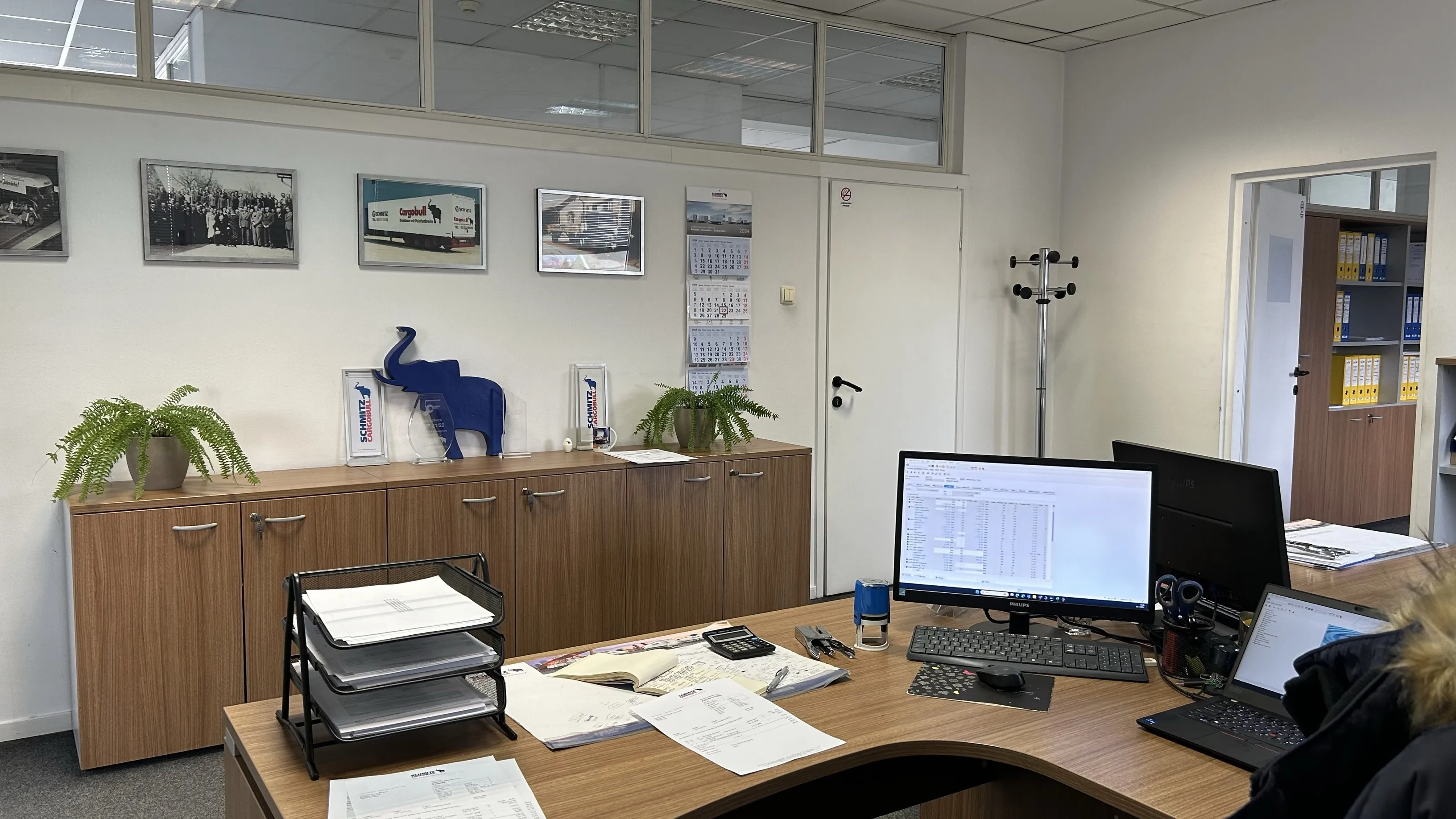 Cozy office vibe at Schmitz Cargobull AG, featuring a desk with computers, a blue elephant decor, and framed truck photos on the wall.