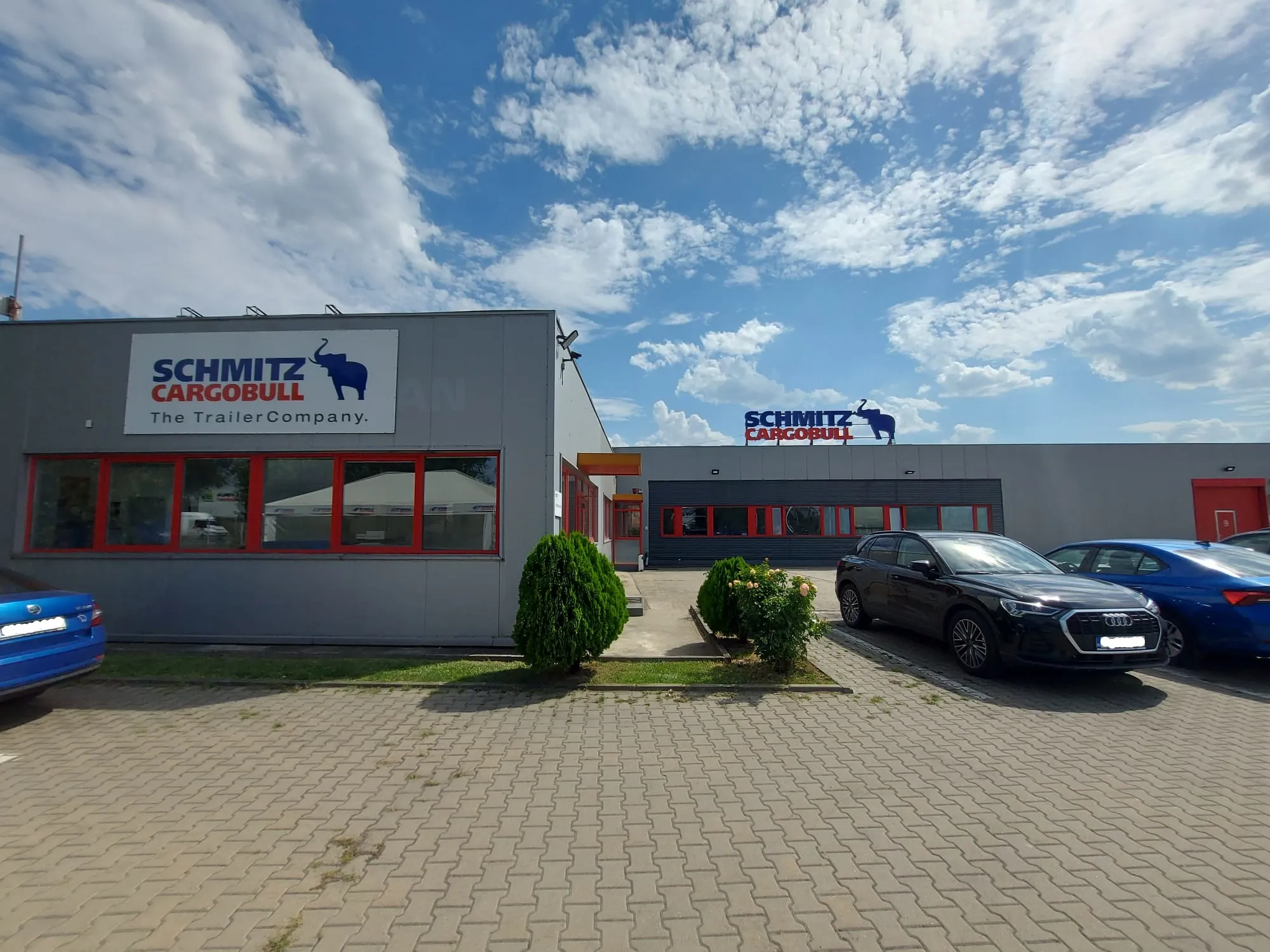 Welcome to Schmitz Cargobull! Check out our sleek office building, surrounded by cars, under a bright, blue sky.