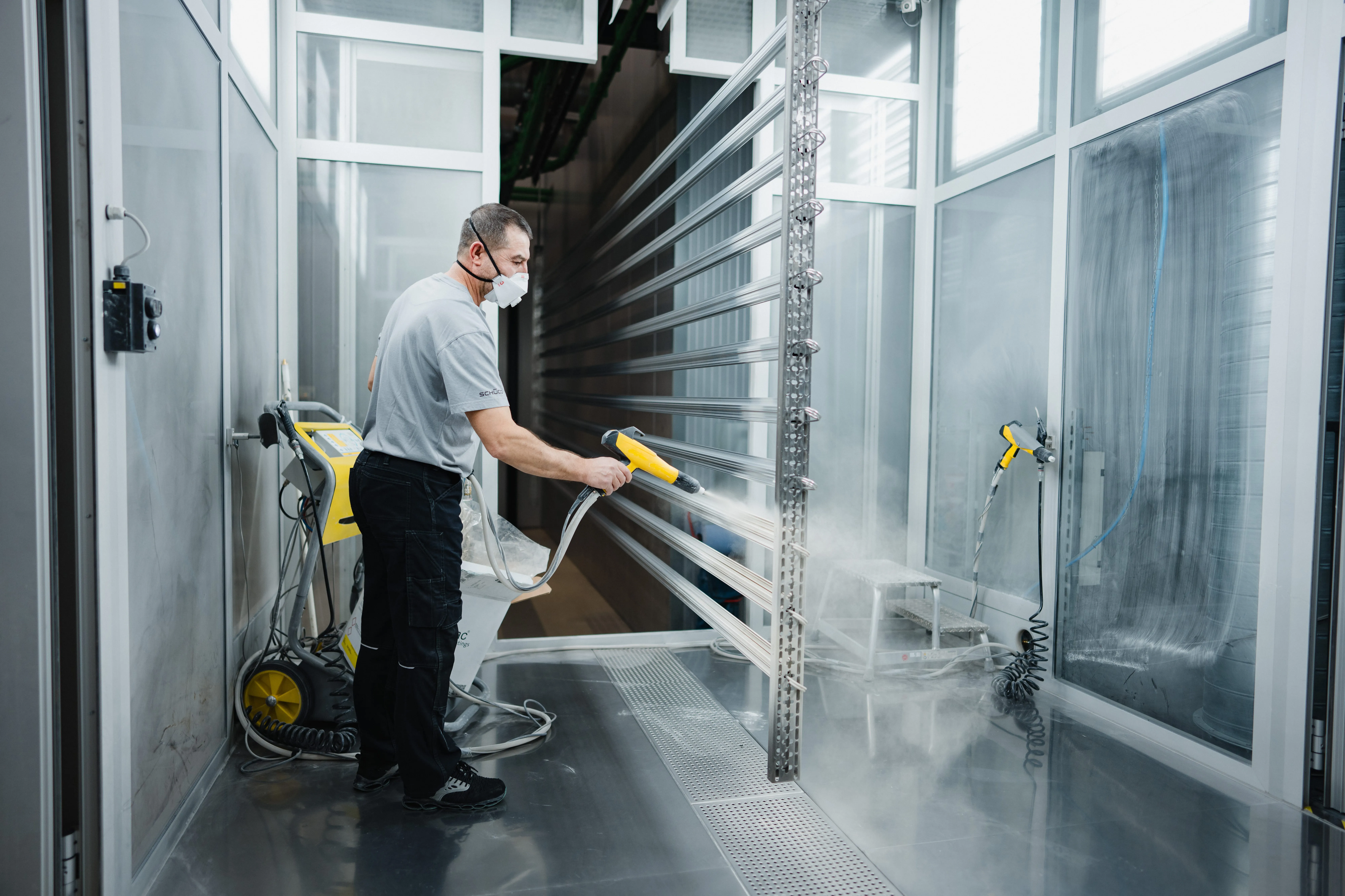 Ein Arbeiter trägt eine Schutzmaske und sprüht ein Metallprofil in einer Lackierkabine bei Schüco International KG.