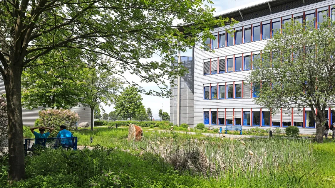 Zwei Mitarbeiter entspannen auf einer Bank im grünen Außenbereich des Schüco-Gebäudes.