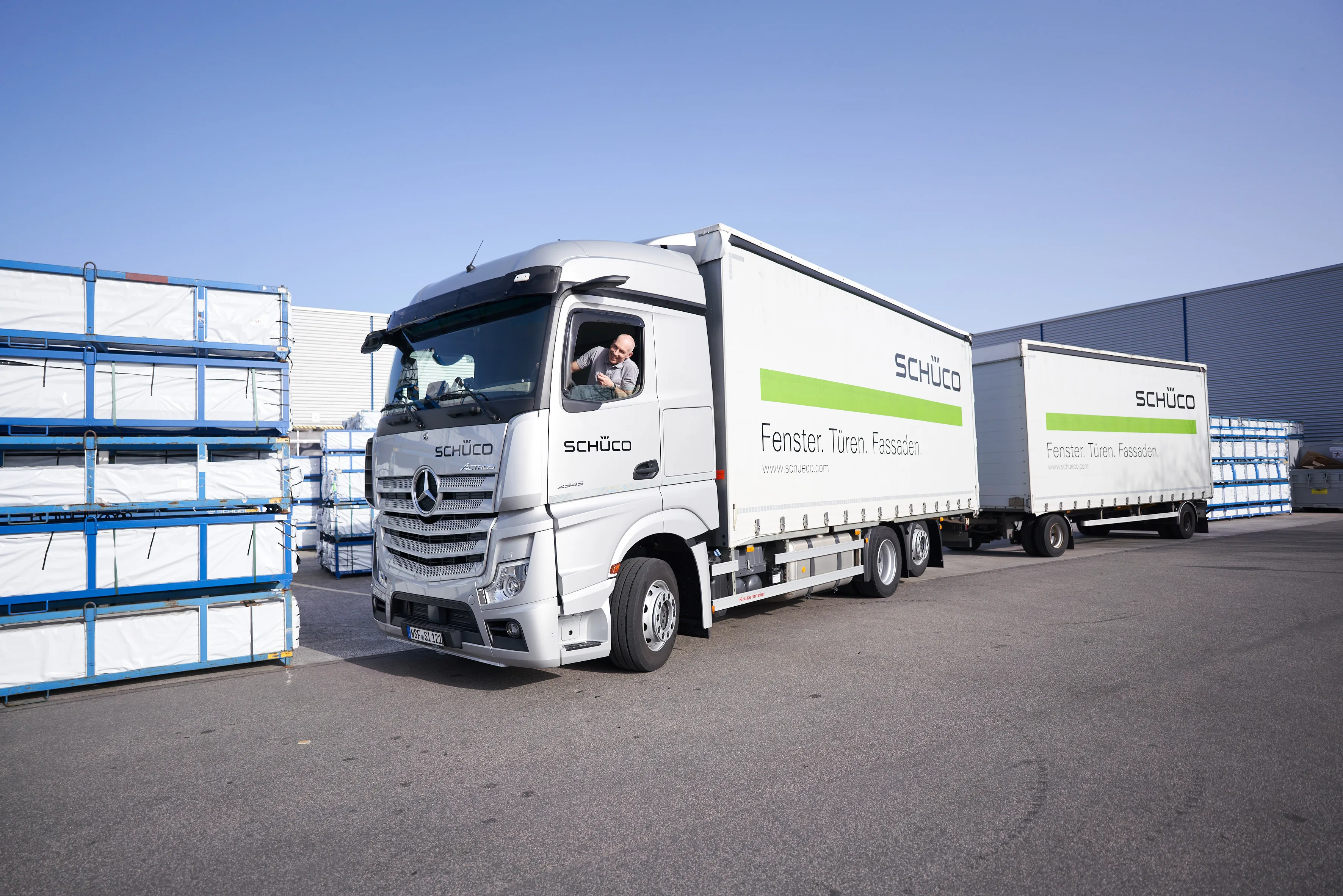 Ein Schüco-LKW-Fahrer grüßt aus dem Fenster, umgeben von gestapelten Baumaterialien auf dem Firmengelände.