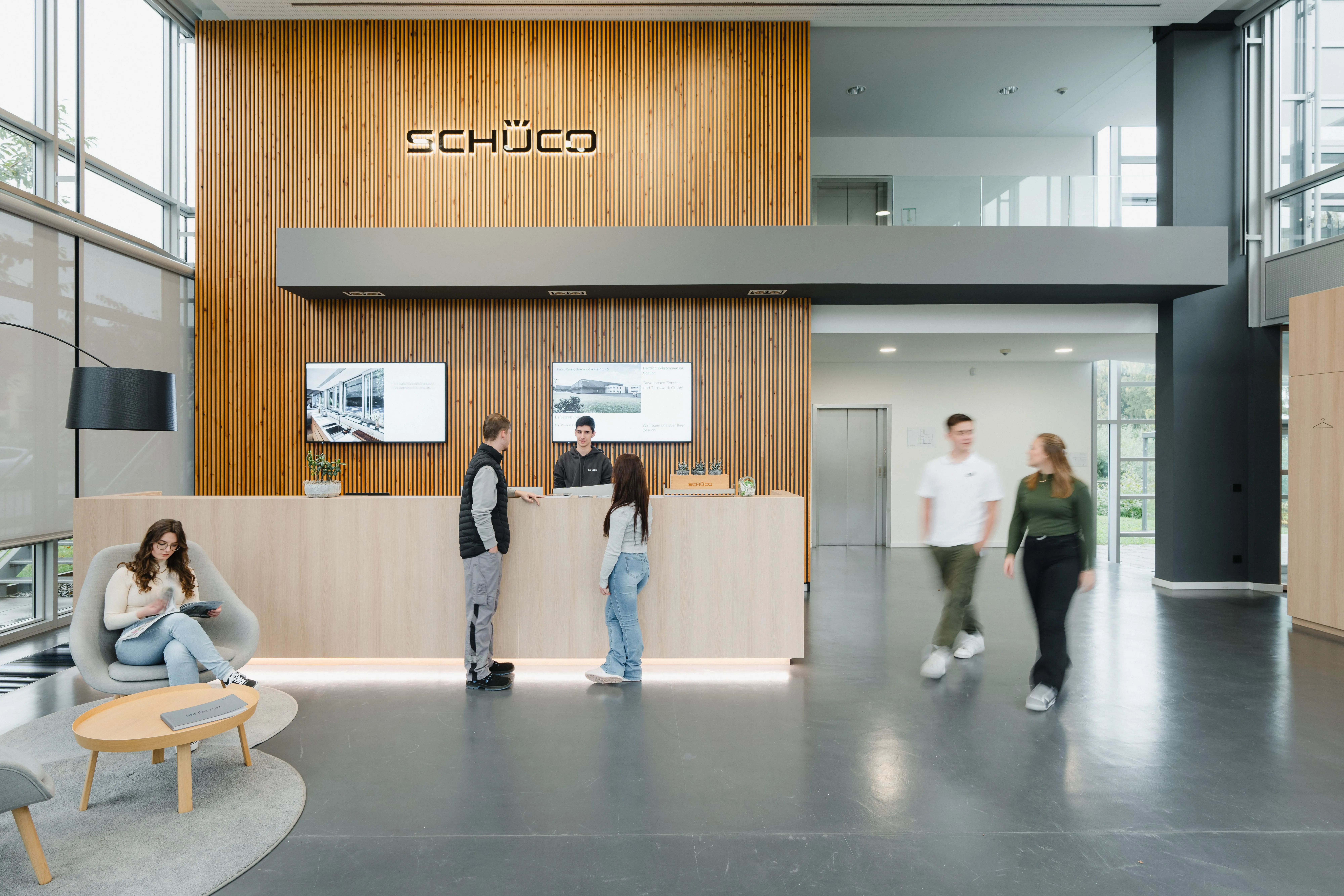 Empfangsbereich bei Schüco International KG mit Personen, die sich unterhalten und warten. Moderne, helle Büroatmosphäre.