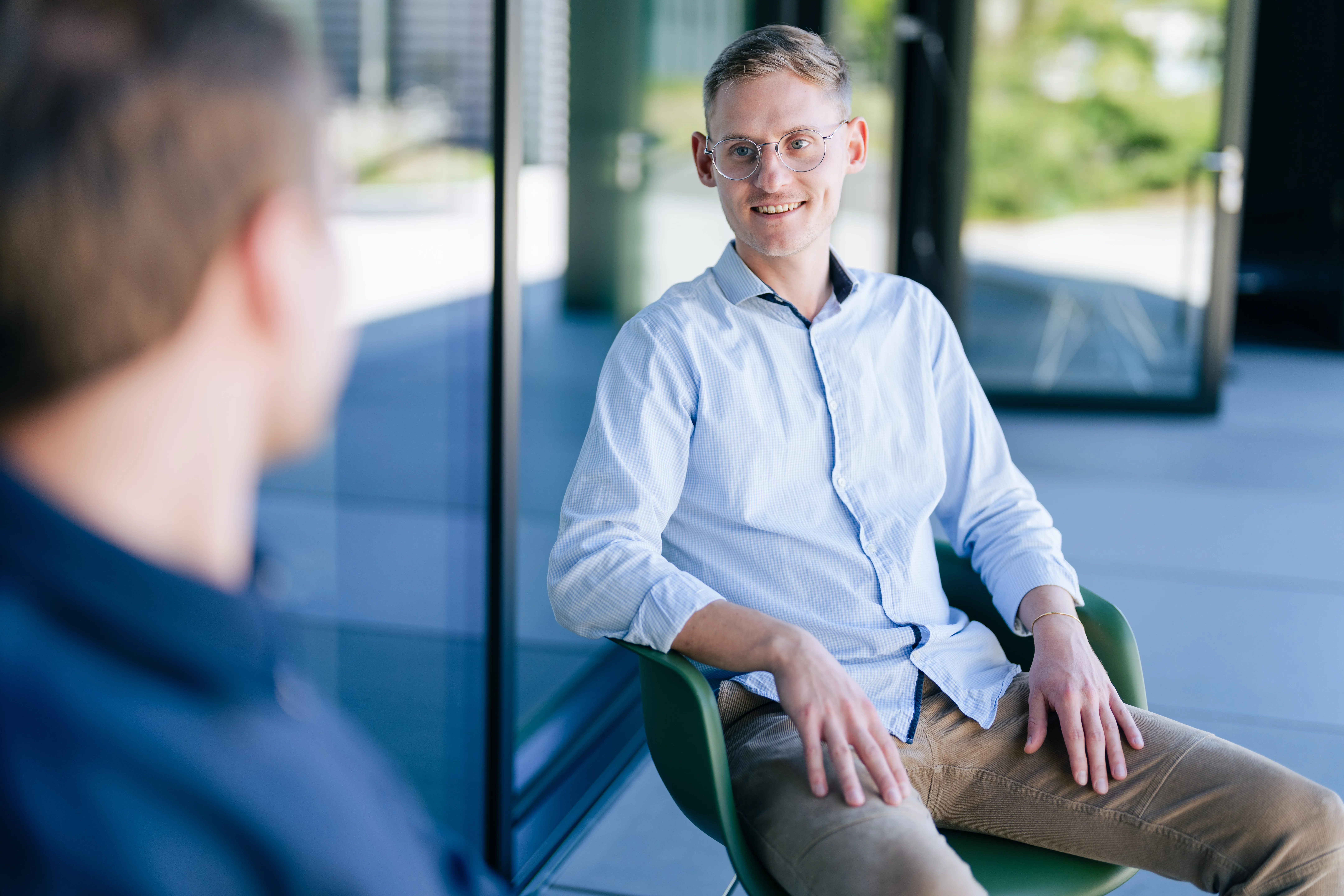 Ein junger Mann in Freizeitkleidung lächelt und sitzt auf einer Terrasse bei Schüco International KG.
