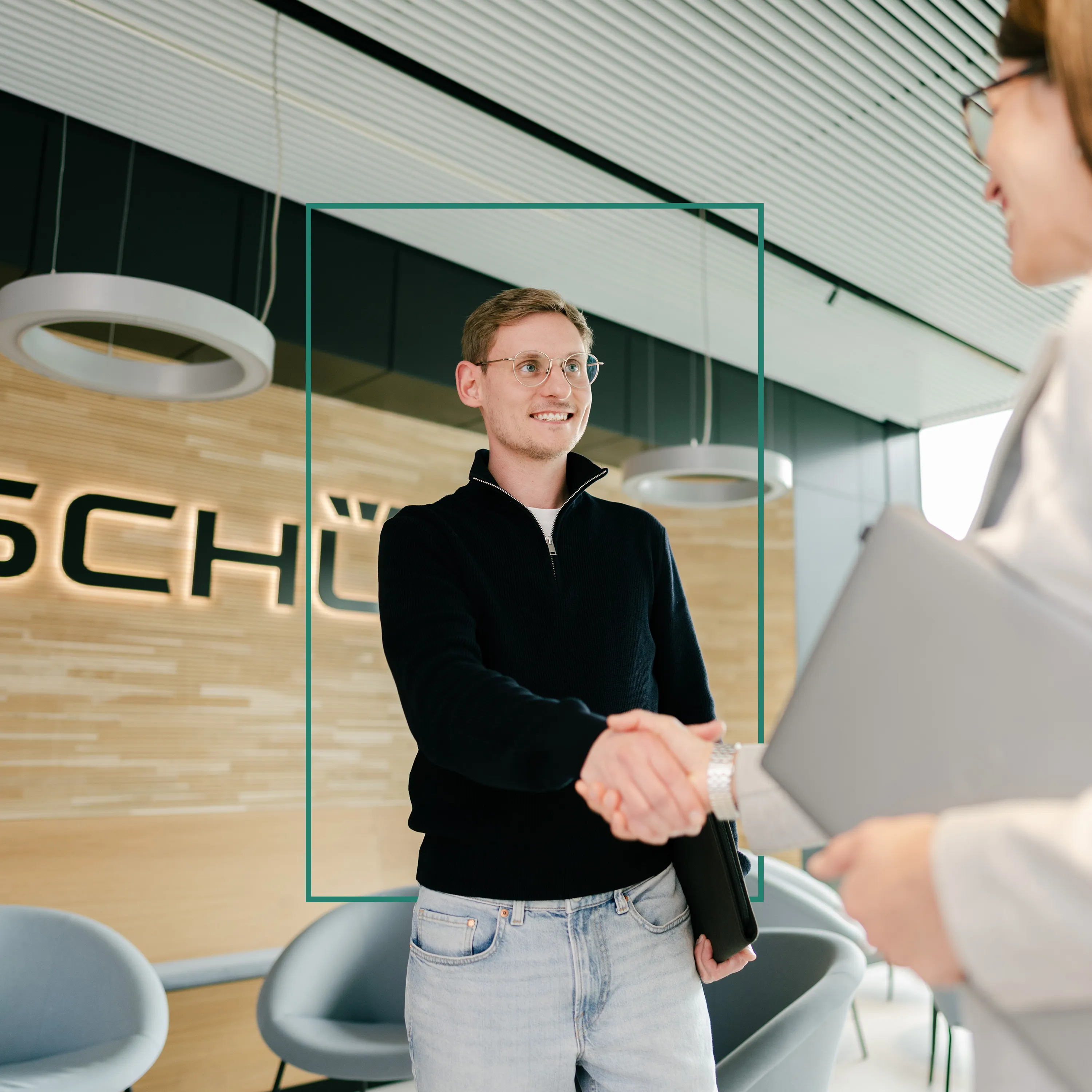 Junger Mann begrüßt Dich mit Handschlag im modernen Schüco-Büro, bereit für Deinen Karrierestart im Bauwesen.