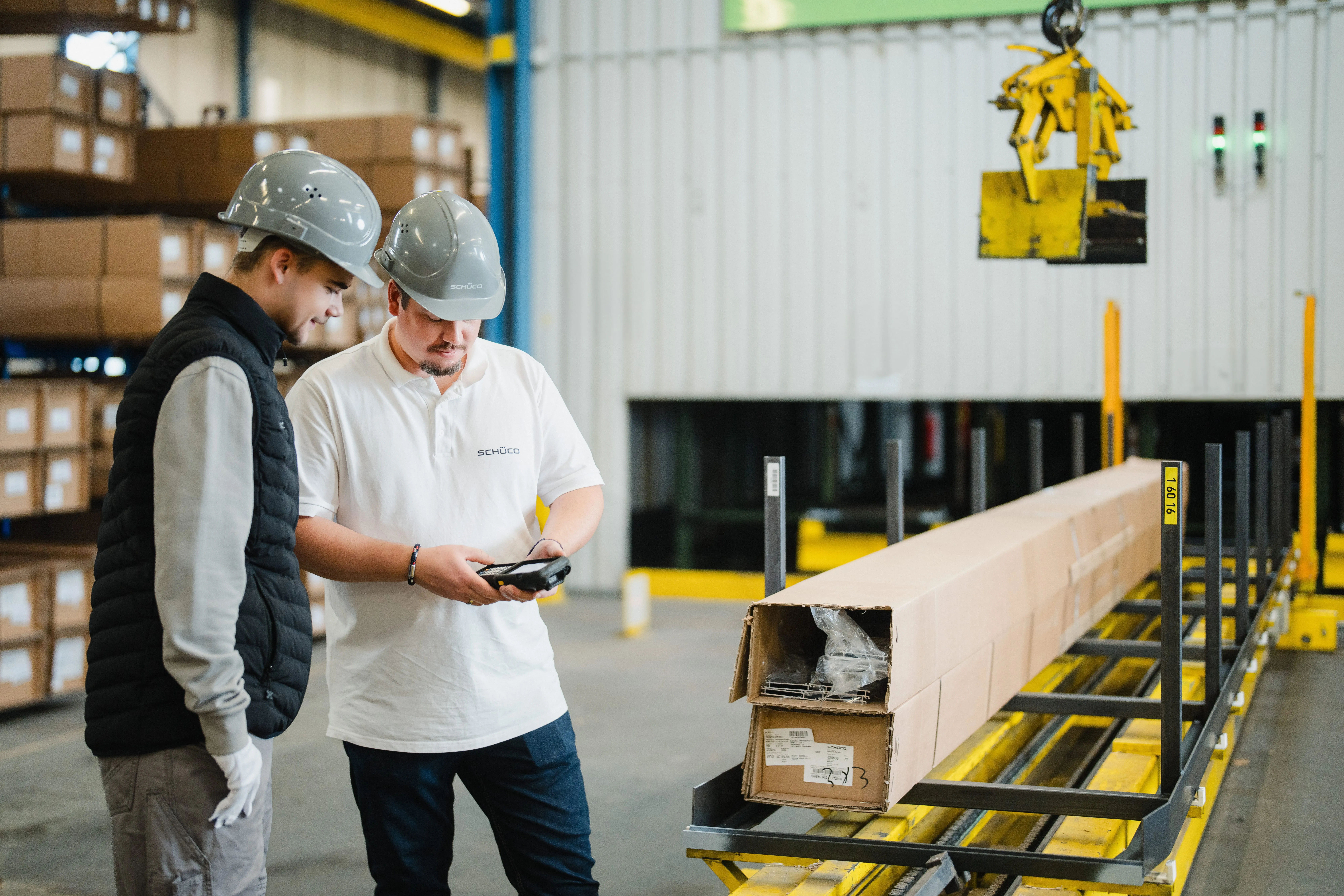 Zwei Arbeiter mit Helmen in einer Lagerhalle von Schüco, die gemeinsam ein Gerät betrachten.
