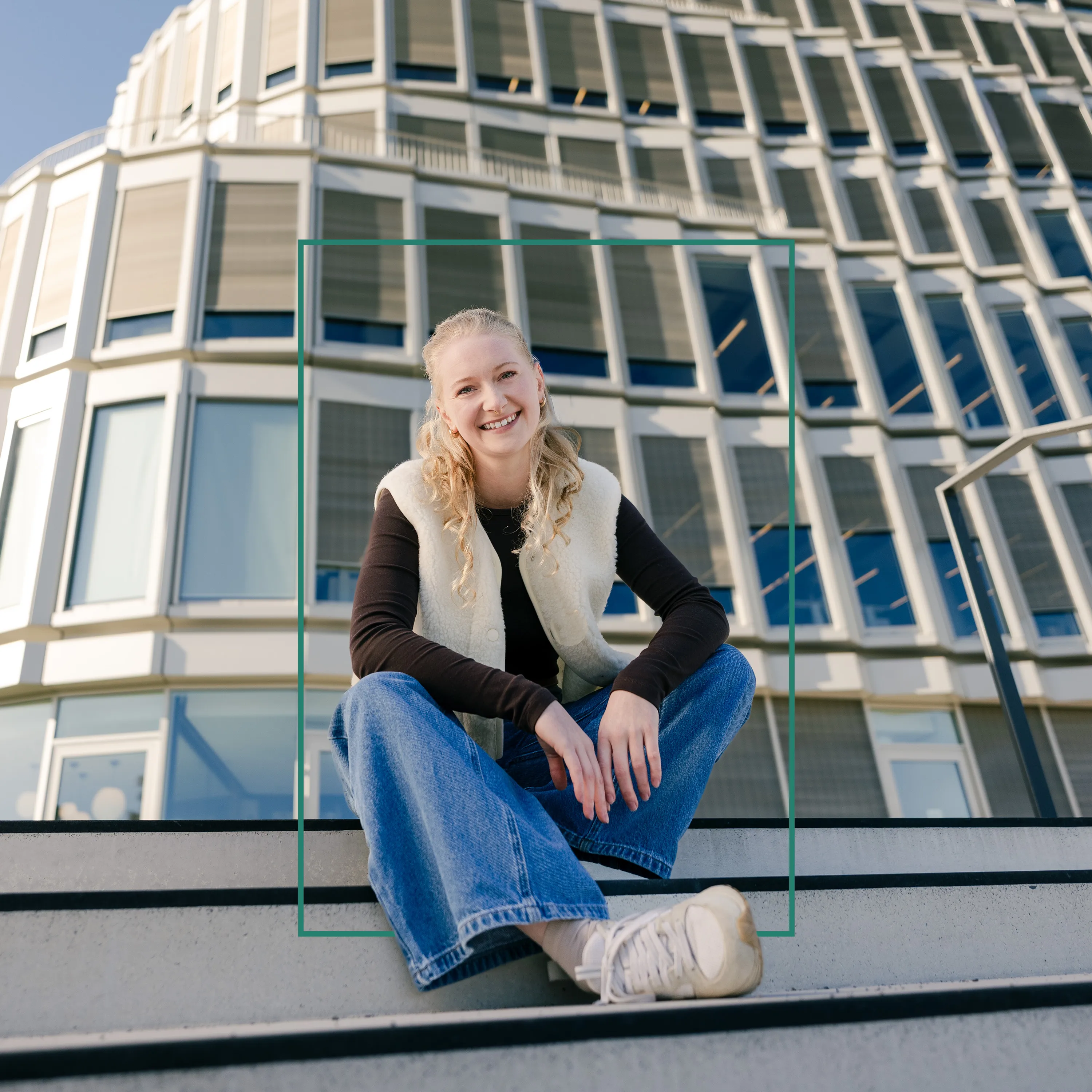 Junge Frau sitzt entspannt vor einem modernen Bürogebäude und lächelt in die Kamera – deine Karriere bei Schüco.