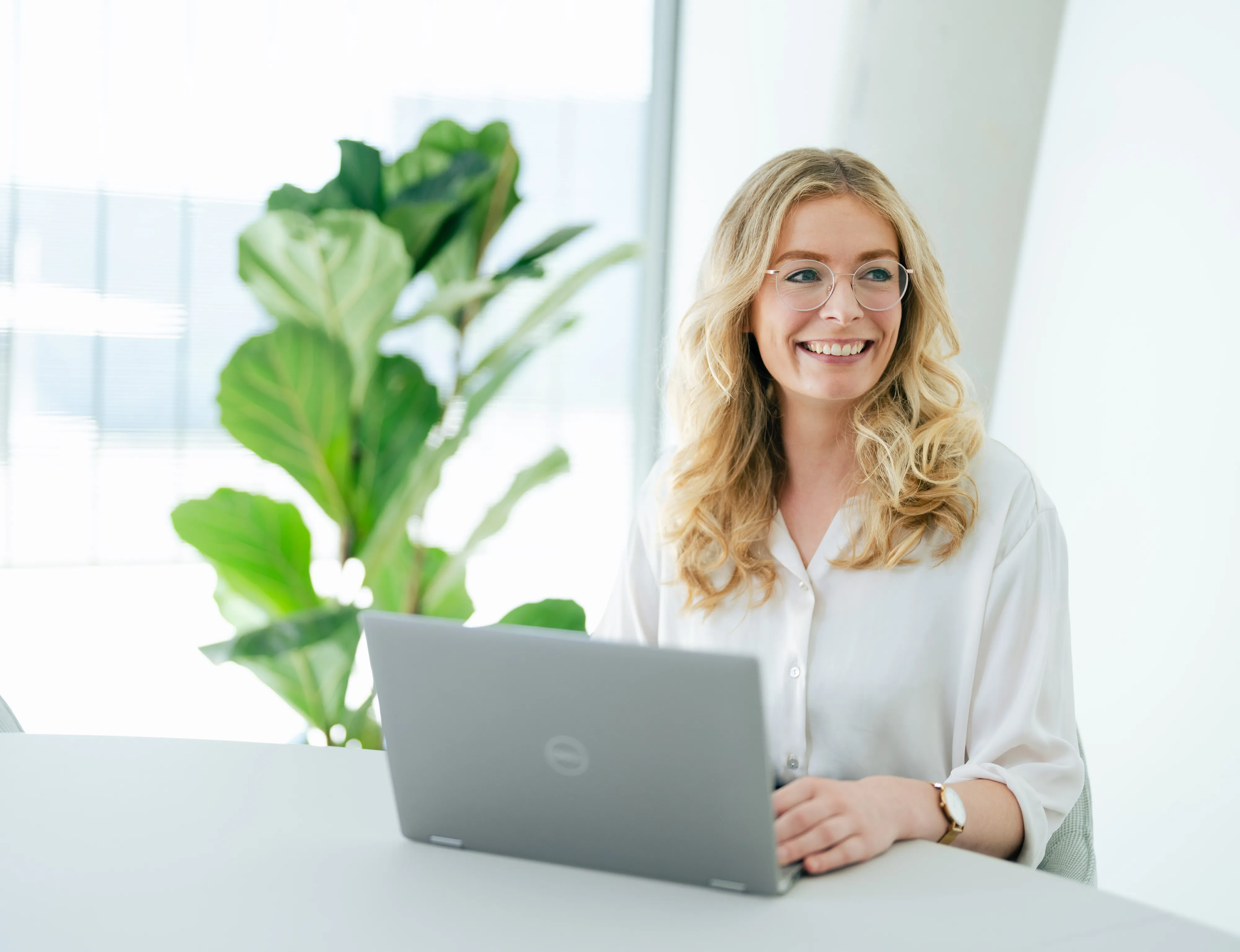 Eine lächelnde Frau im Büro arbeitet an einem Laptop, im Hintergrund eine grüne Pflanze.