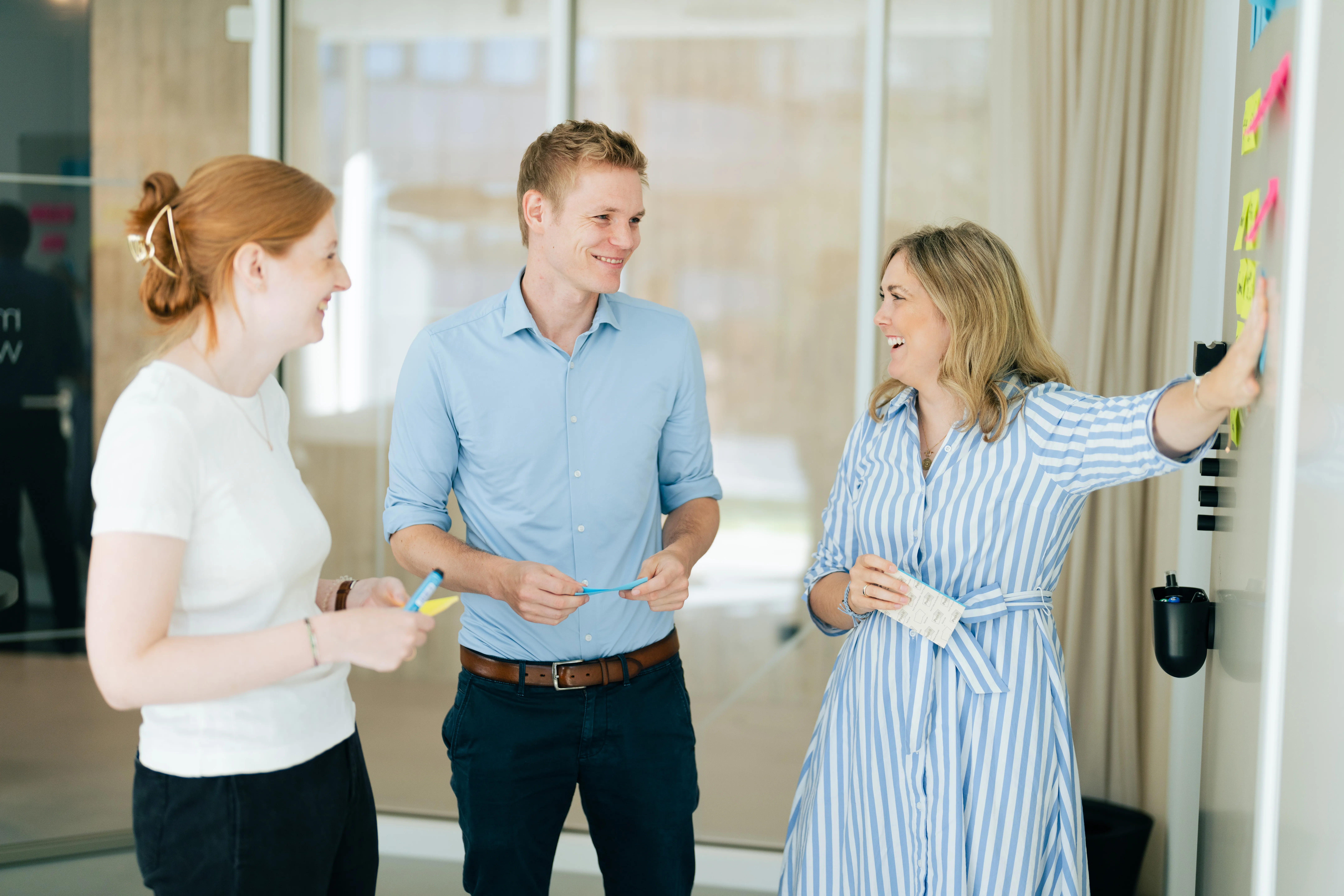 Drei Kollegen diskutieren fröhlich vor einem Whiteboard mit Haftnotizen in einem modernen Büro bei Schüco International KG.