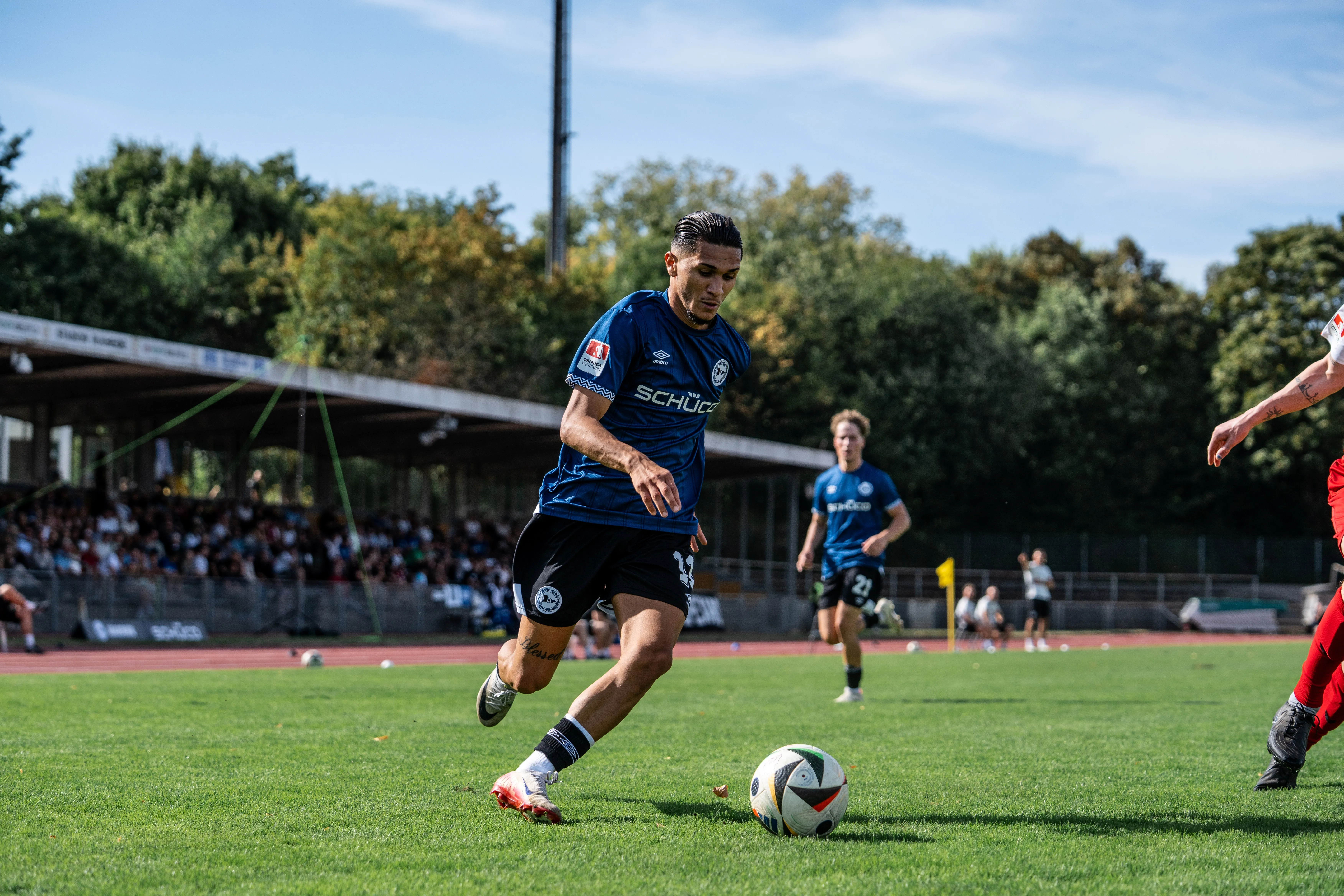 Ein Fußballspieler im Schüco-Trikot dribbelt den Ball über ein Fußballfeld, während Zuschauer im Hintergrund zuschauen.
