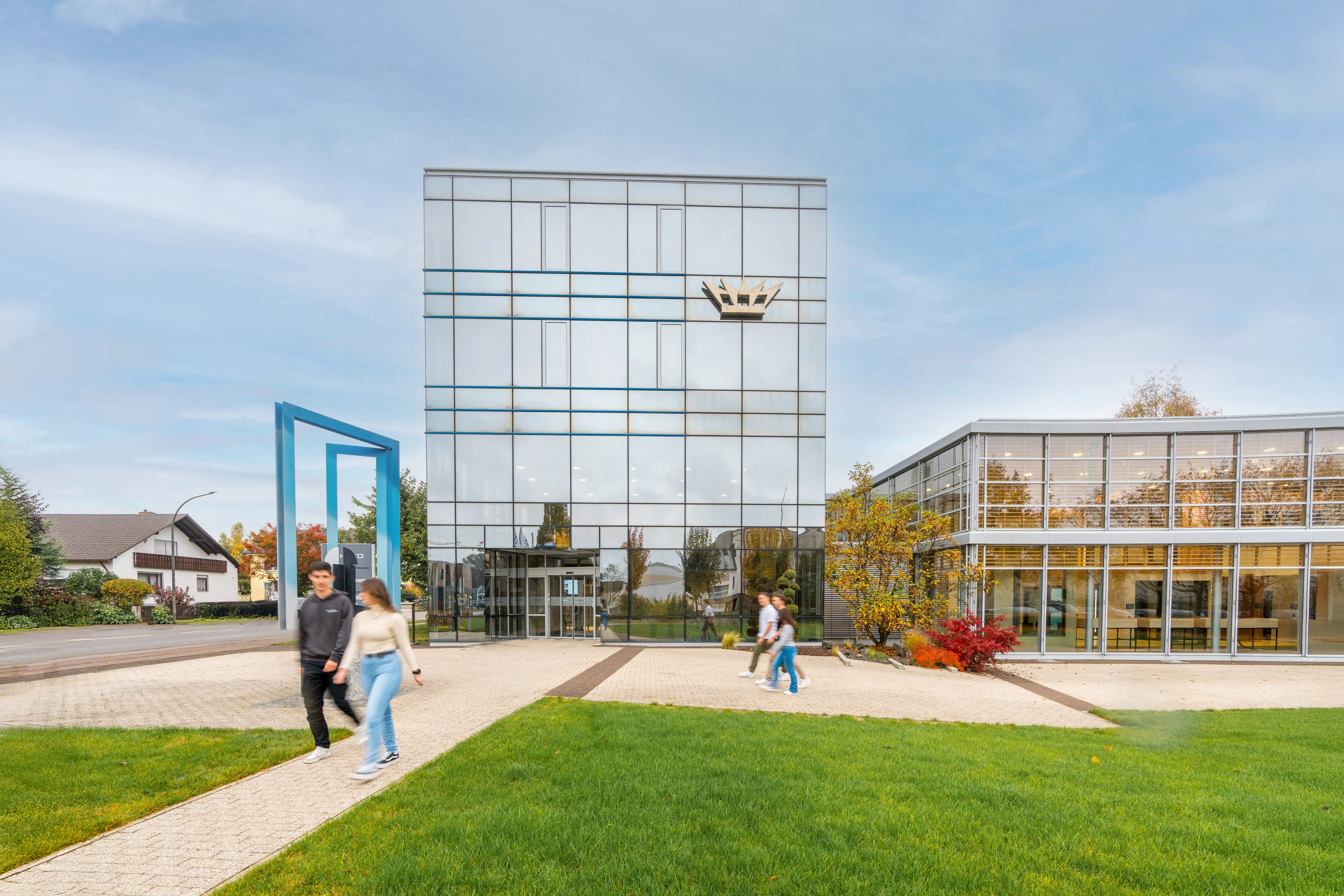 Moderne Bürogebäude von Schüco International KG mit Glasfassaden und Personen, die den Eingang passieren, in einer grünen Umgebung.