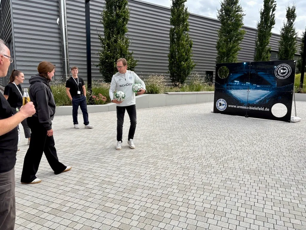 Eine Gruppe von Personen steht im Freien vor einer Torwand mit Schüco-Logo.