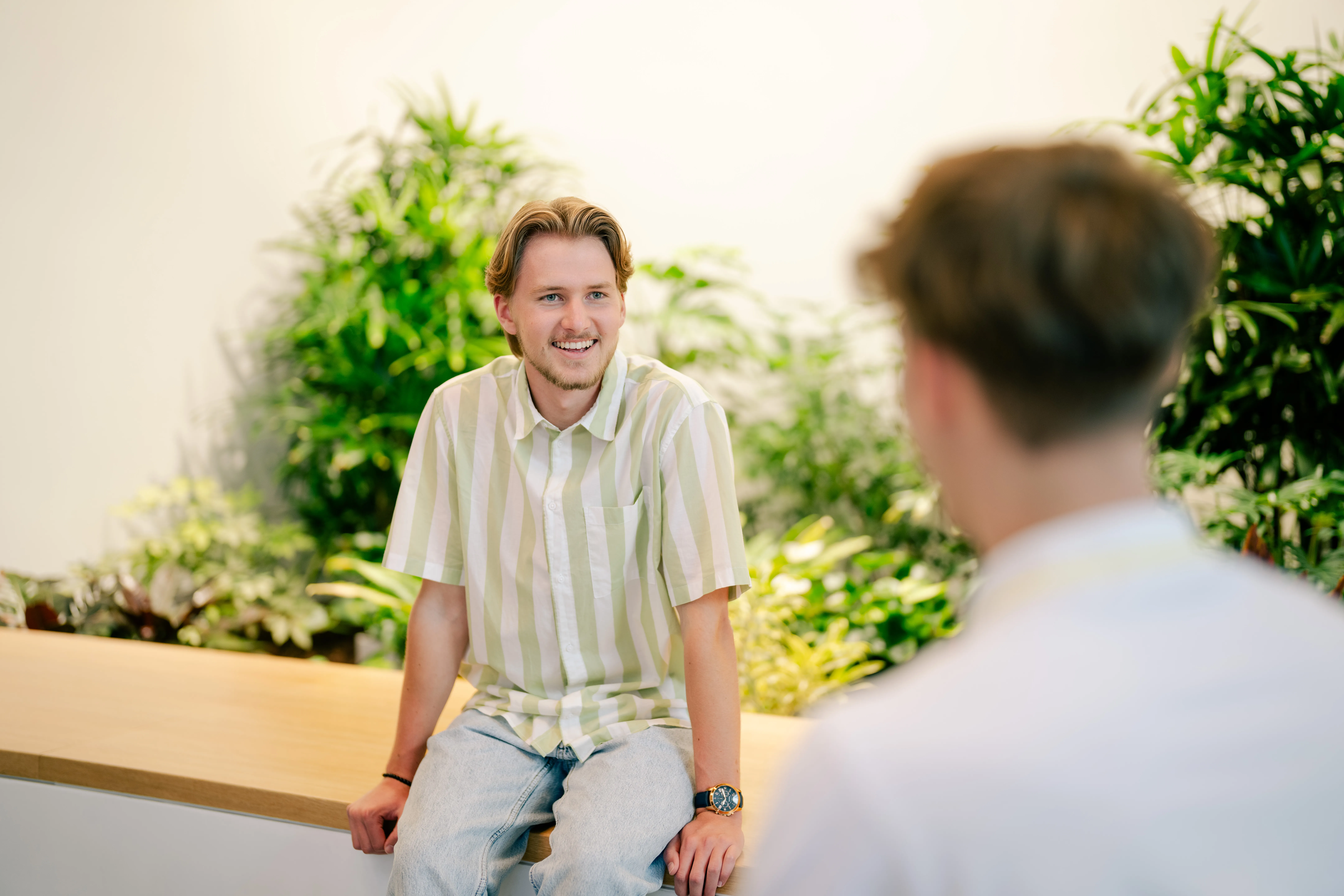 Ein junger Mann im Gespräch mit einem Kollegen in einem modernen Büro mit Pflanzenhintergrund bei Schüco International KG.
