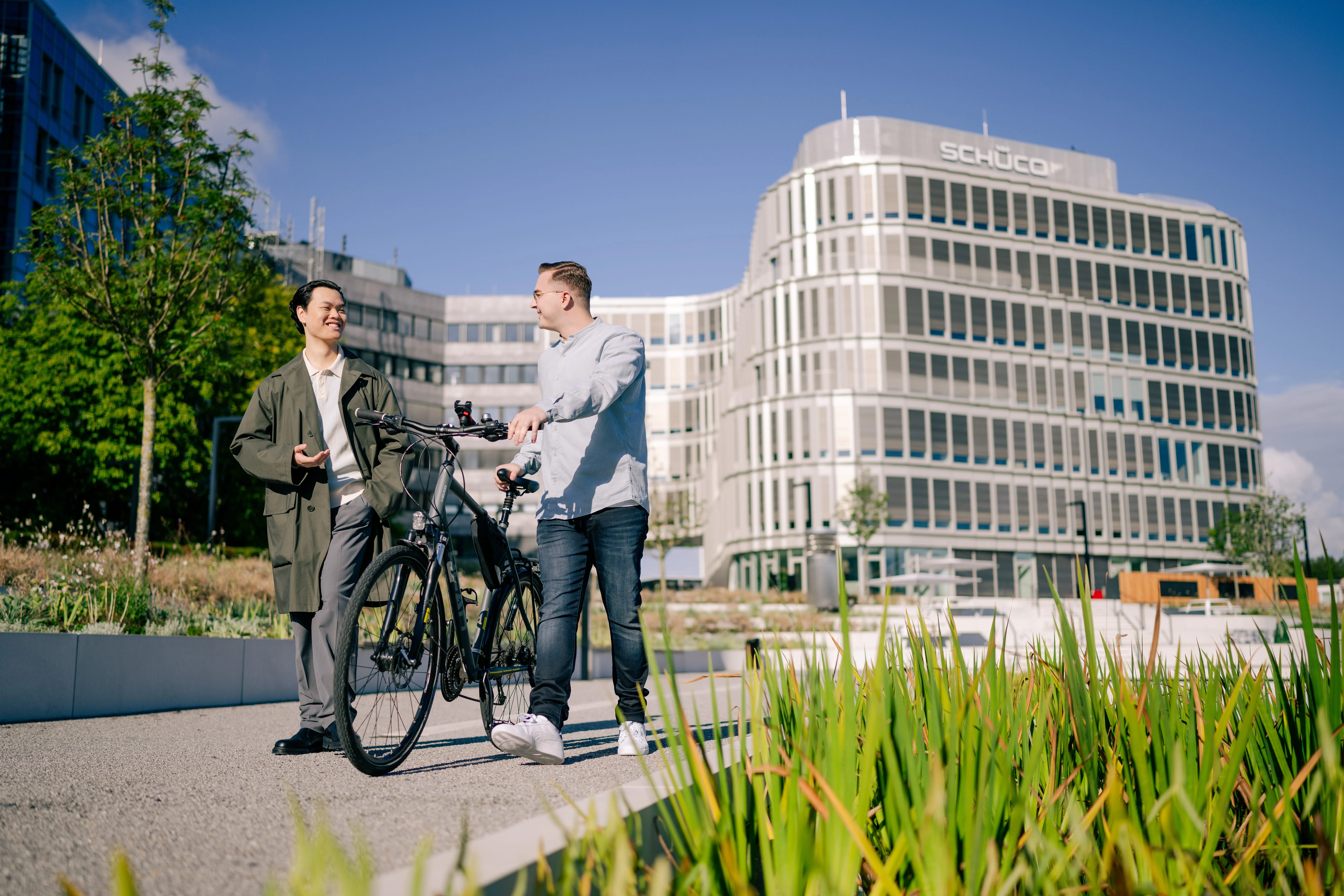 Zwei Personen unterhalten sich vor dem Schüco-Gebäude, eine mit einem Fahrrad. Ideal für die Schüco Karriere-Website.