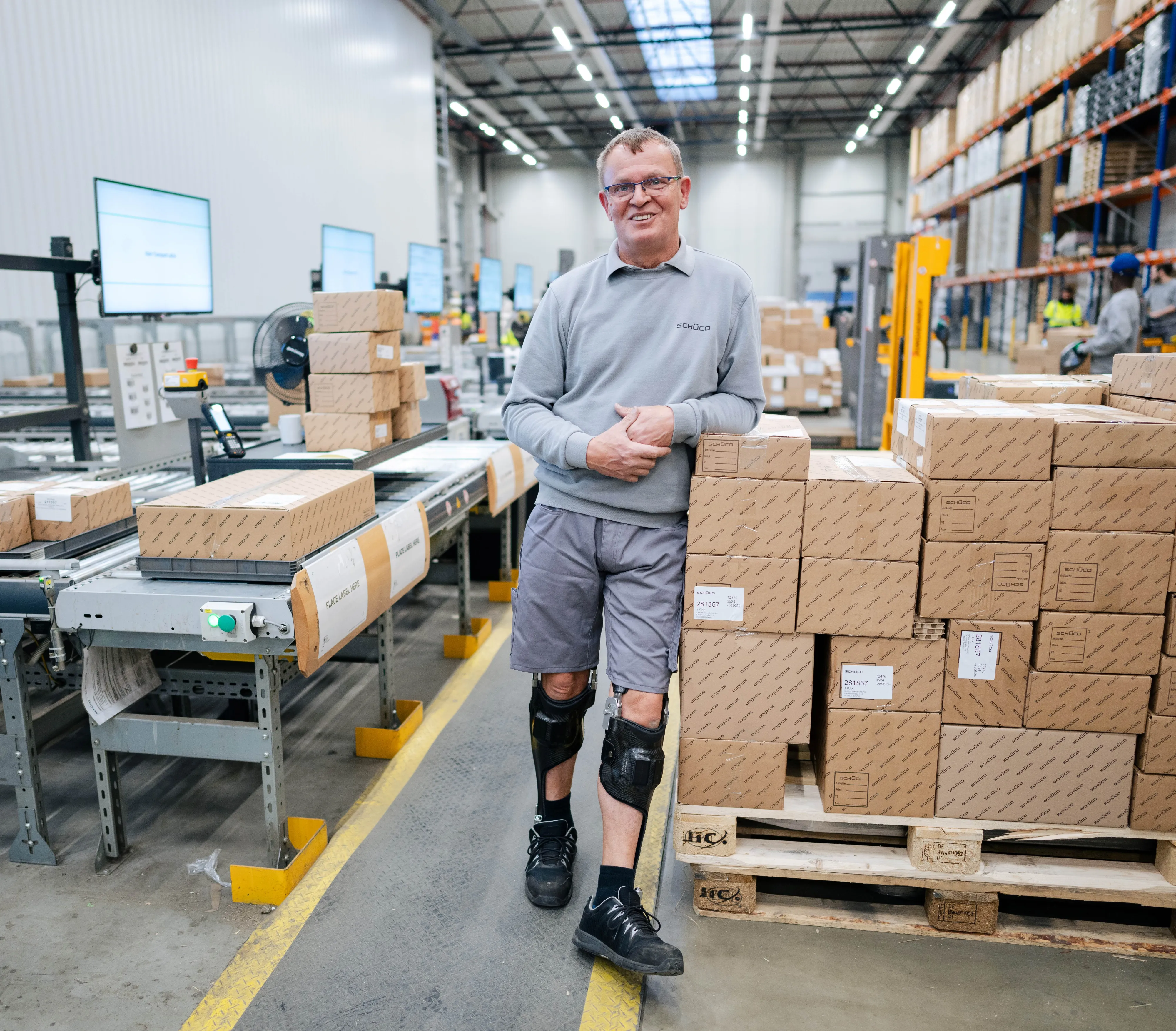 Mitarbeiter in Schüco-Arbeitskleidung lehnt an Paketstapel im modernen Lager, umgeben von Kartons und Förderband.