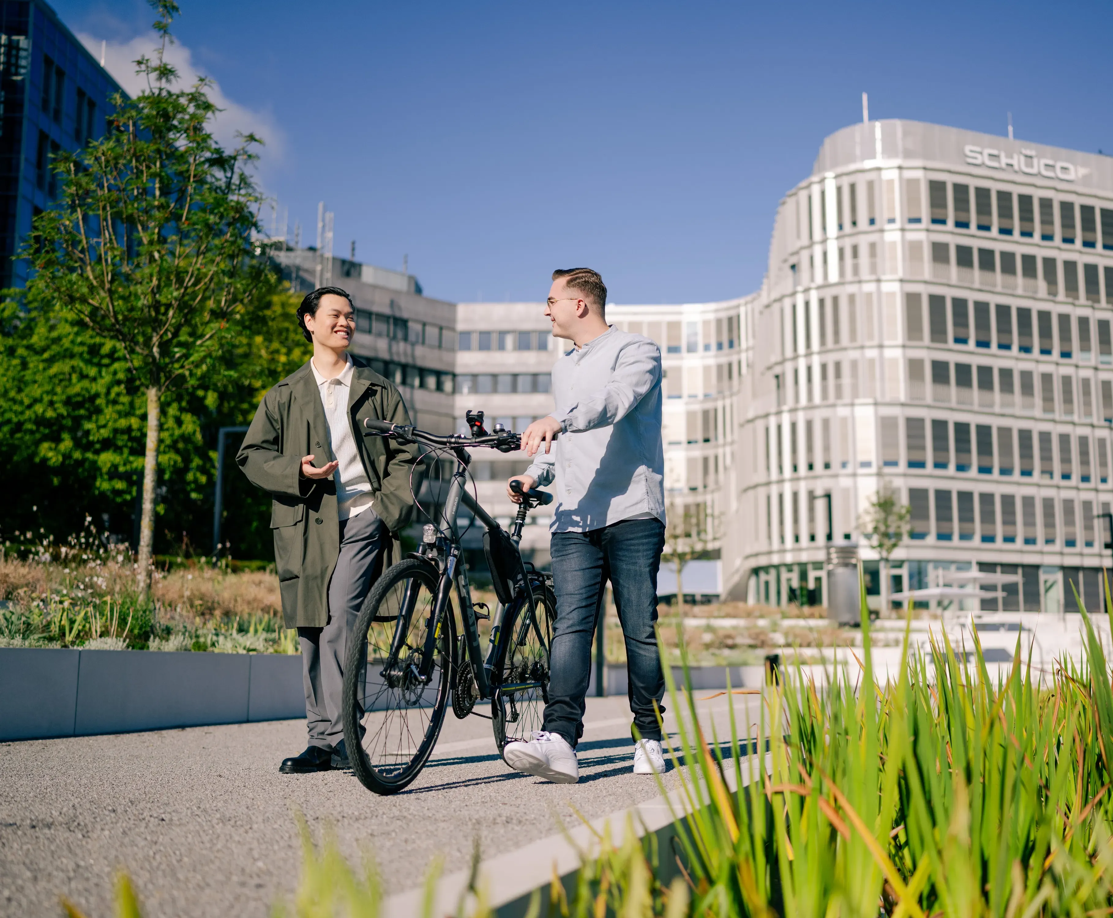 Zwei Kollegen unterhalten sich fröhlich neben einem Fahrrad vor dem Schüco-Gebäude.