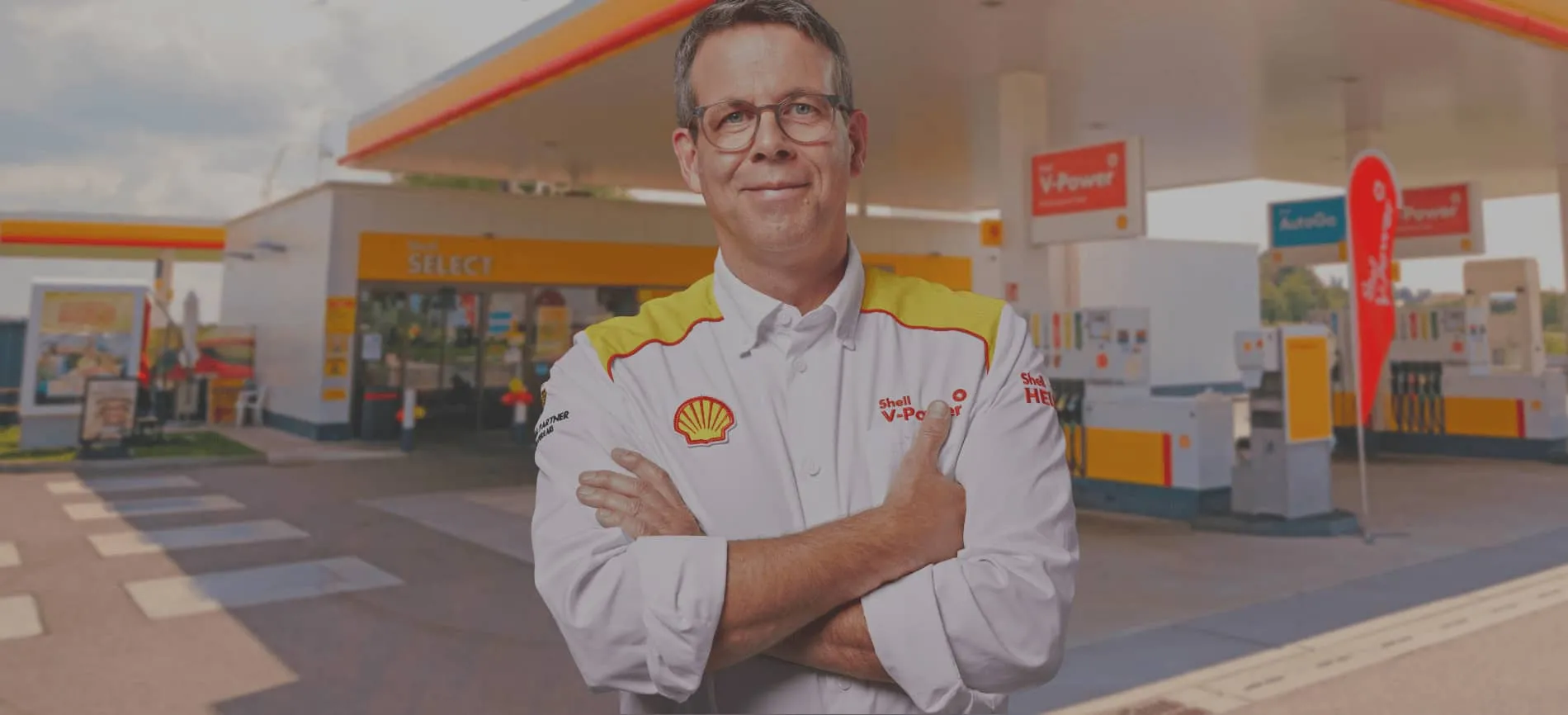 Ein Shell-Mitarbeiter steht selbstbewusst vor einer Shell-Tankstelle und begrüßt Dich als potenziellen neuen Kollegen.