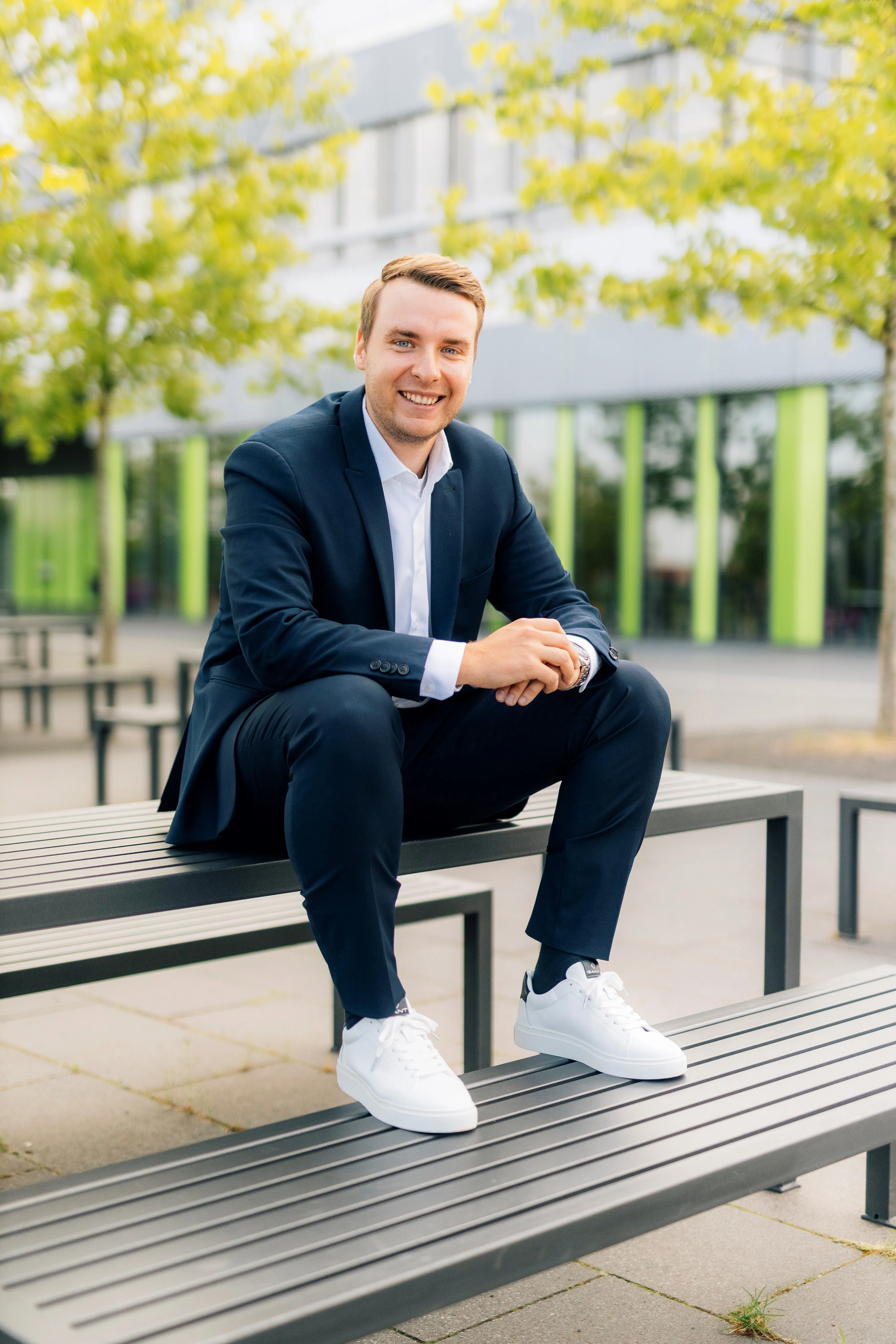 Ein lächelnder Mann in einem Anzug sitzt draußen auf einer Bank vor einem modernen Bürogebäude.