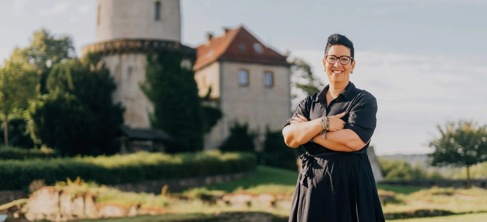 Eine lächelnde Person steht mit verschränkten Armen vor der Sparrenburg in Bielefeld und blickt selbstbewusst in die Kamera.