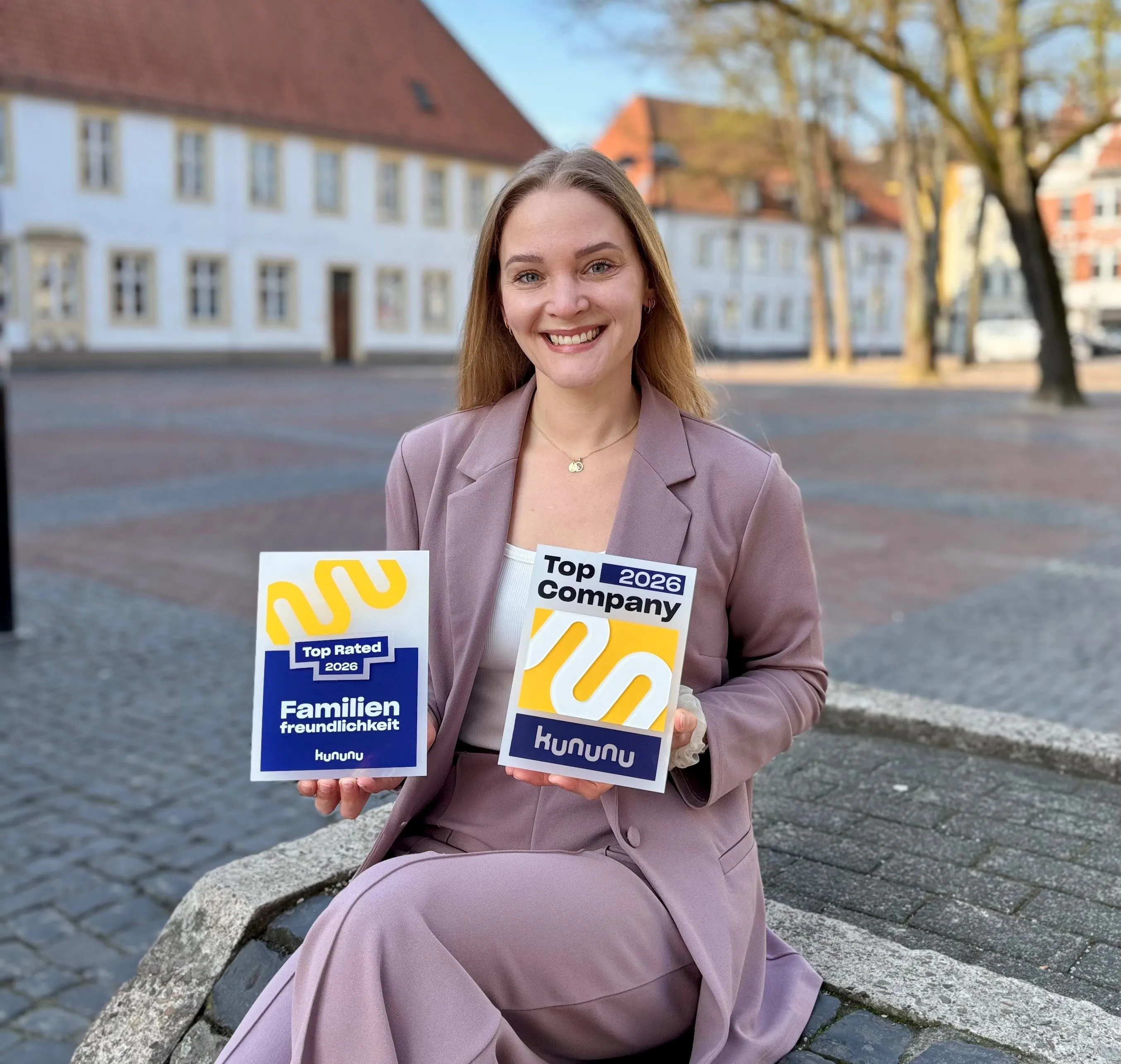 Eine Frau im Business-Outfit sitzt draußen vor einem modernen Gebäude und hält ein "Top Company 2023"-Schild von kununu.