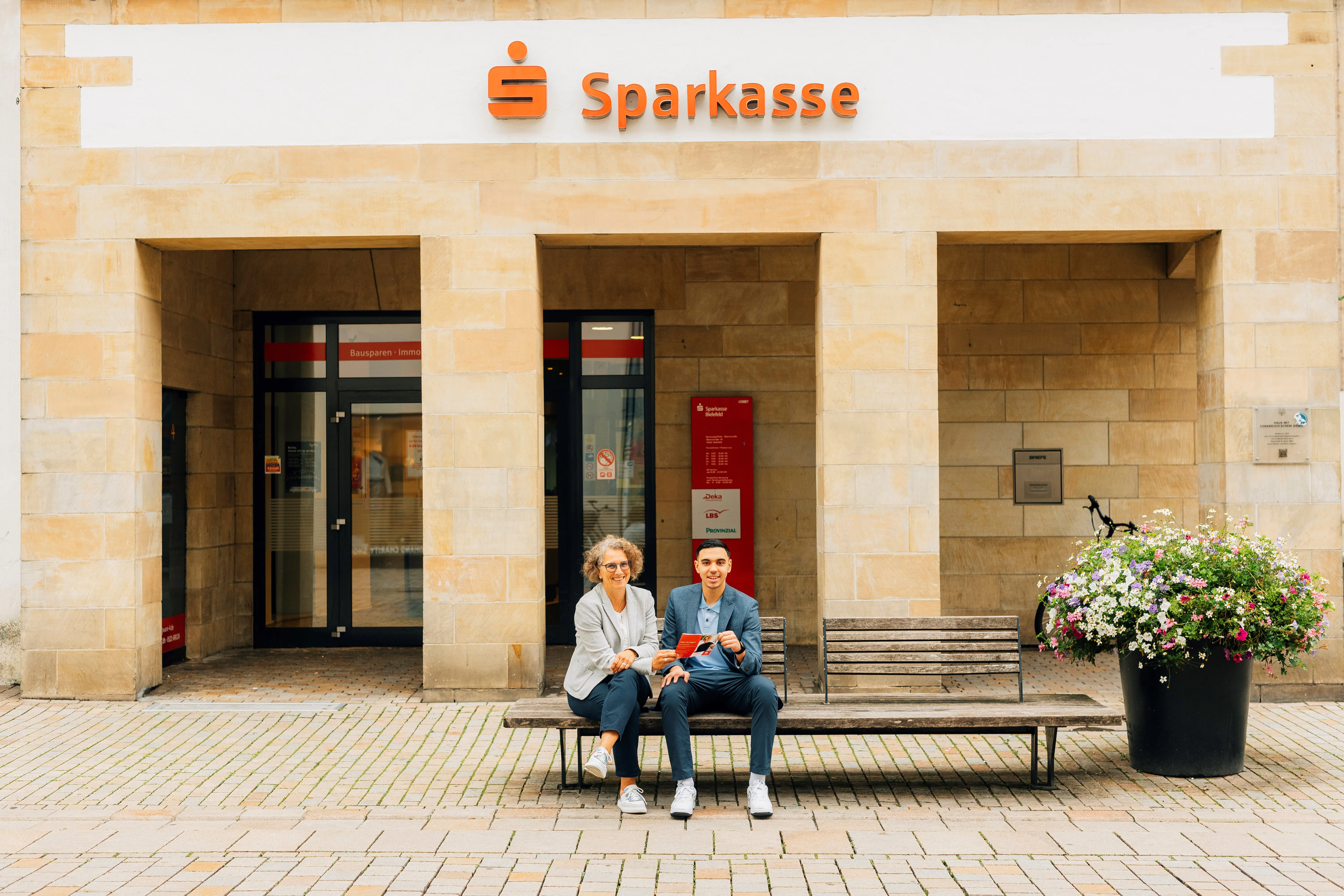 Zwei Personen sitzen auf einer Bank vor der Sparkasse Bielefeld und halten Flyer.