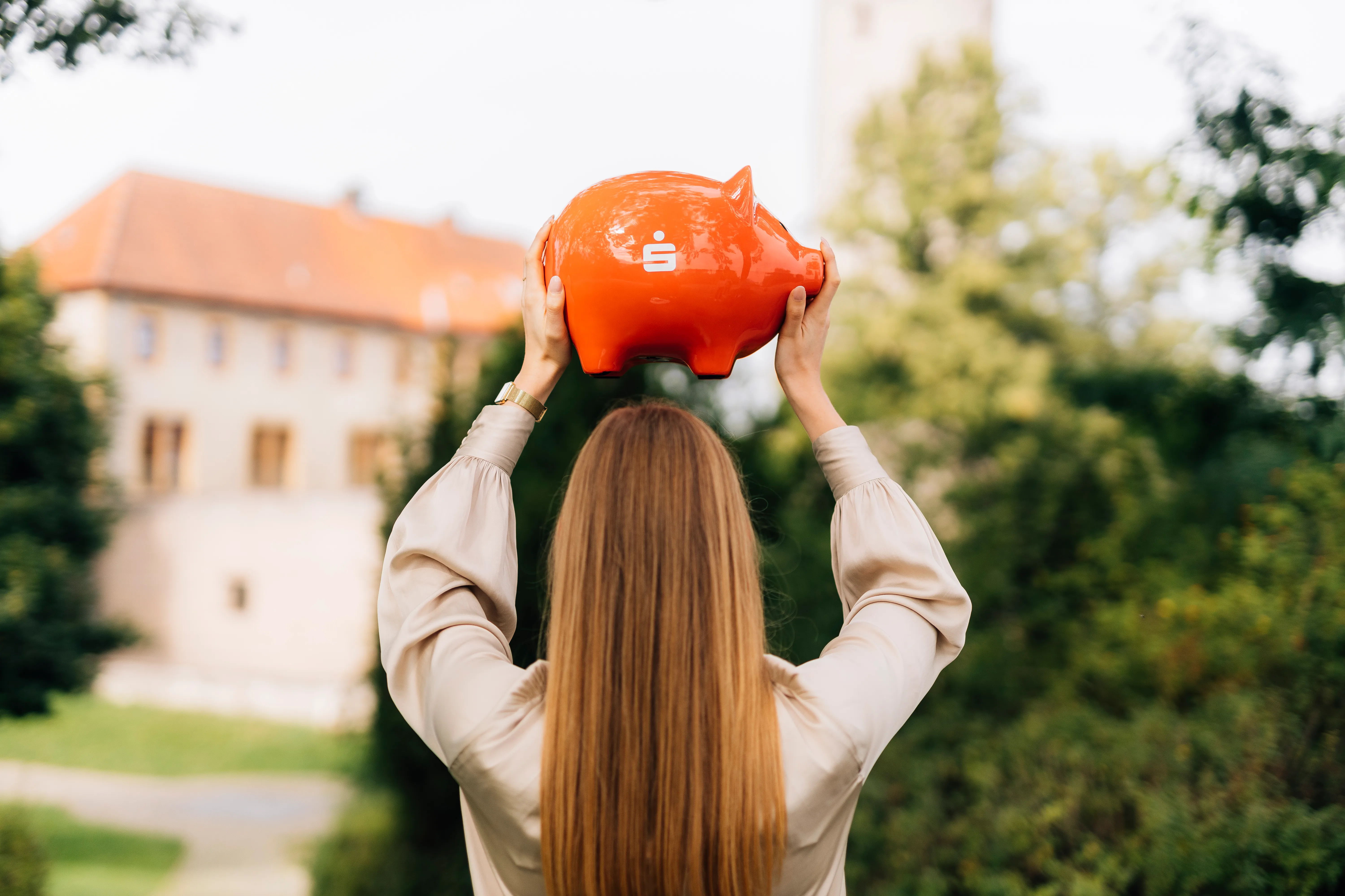 Eine Frau hebt ein rotes Sparschwein mit Sparkasse-Logo in die Luft, im Hintergrund ein Gebäude und Bäume.