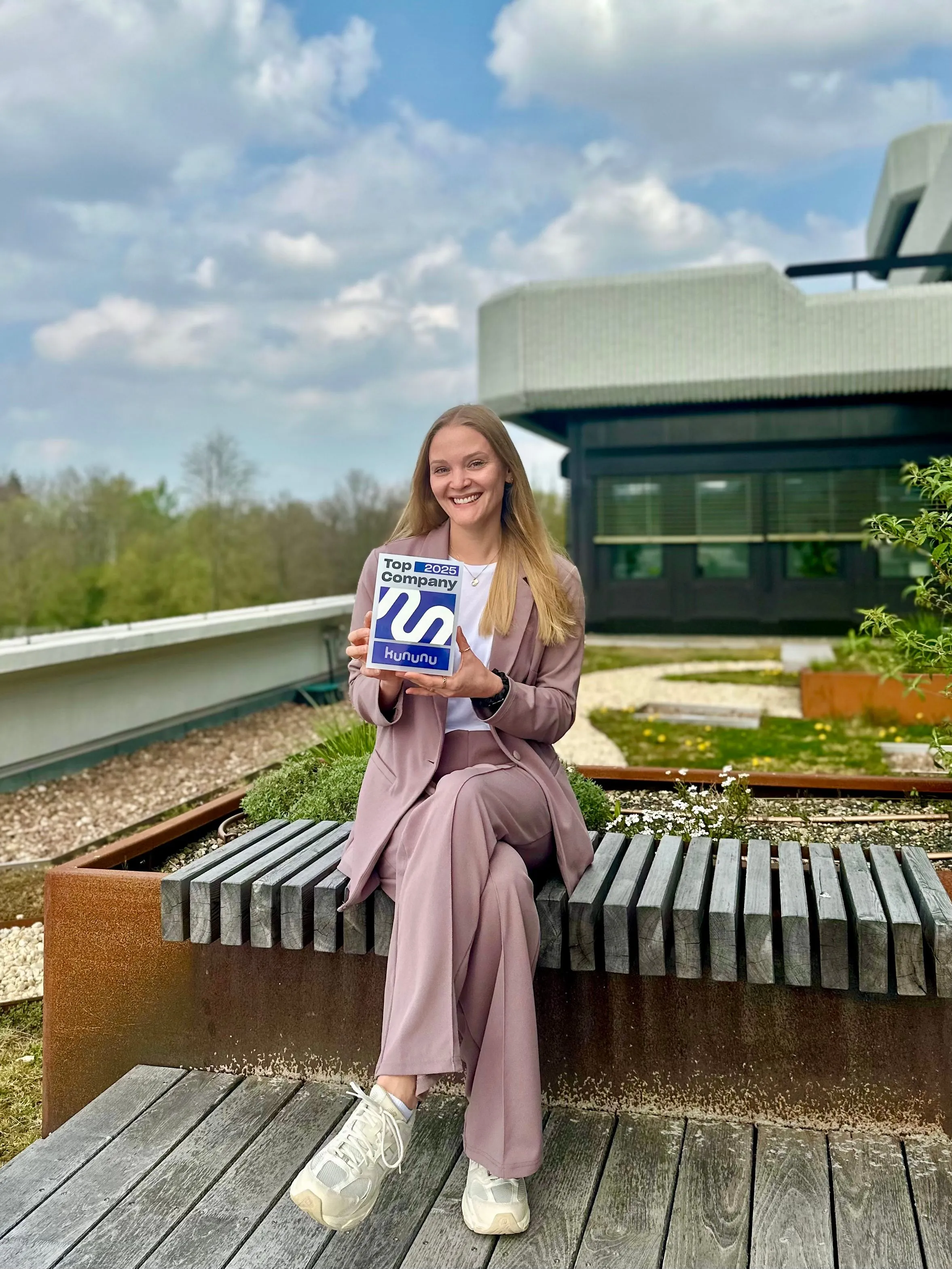 Eine lächelnde Frau im Anzug hält eine "Top Company 2025"-Auszeichnung von Kununu. Sie sitzt lässig draußen auf einer Bank.