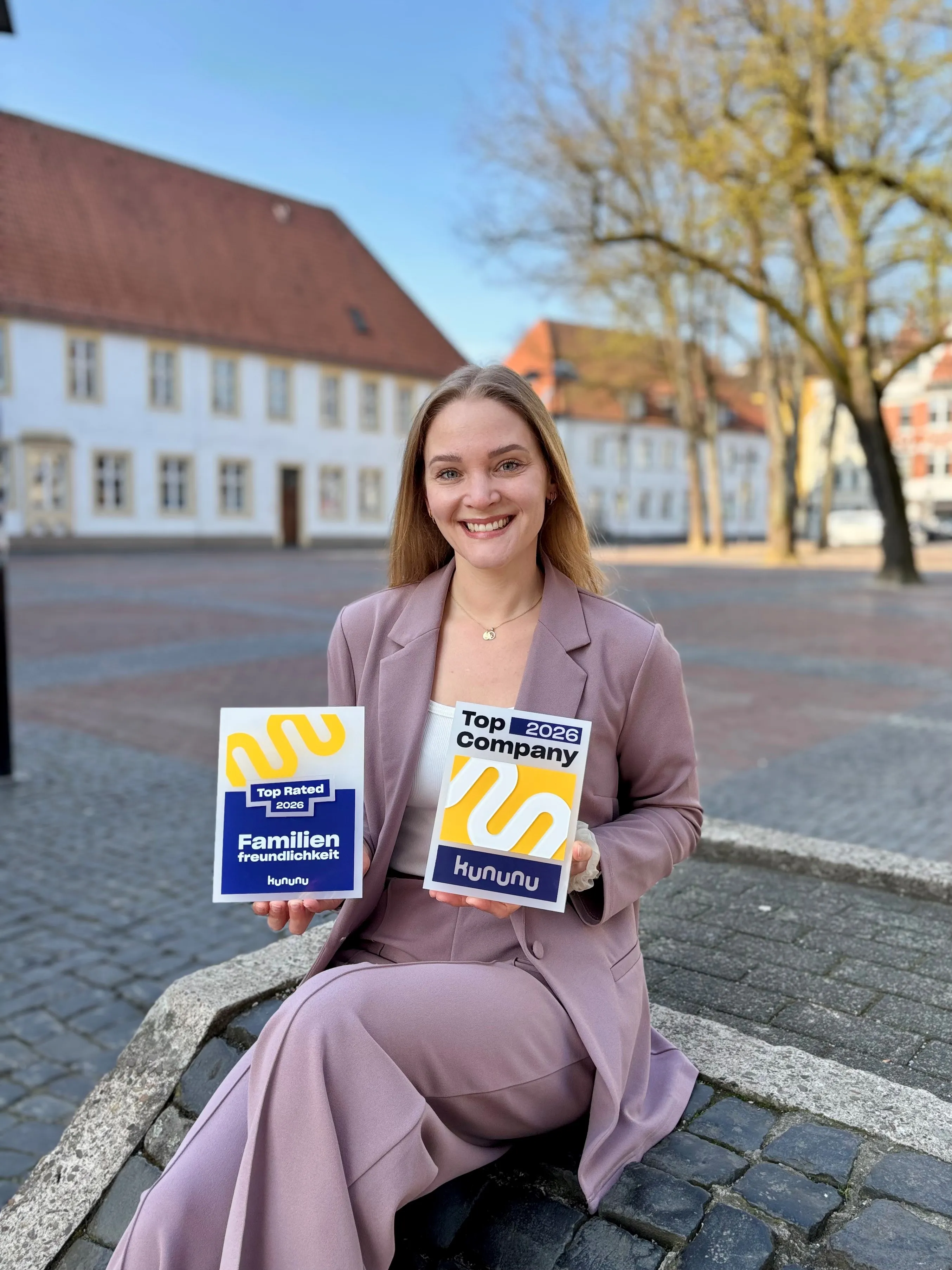 Eine lächelnde Frau im Anzug hält eine "Top Company 2025"-Auszeichnung von Kununu. Sie sitzt lässig draußen auf einer Bank.