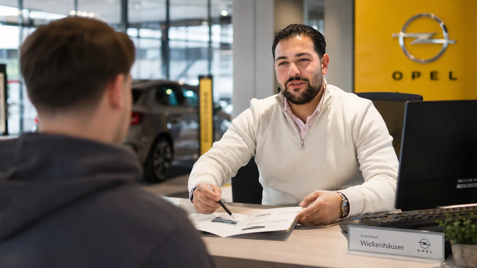 Du siehst einen Opel-Berater, der einem Kunden im Autohaus ein Angebot erklärt und freundlich berät.