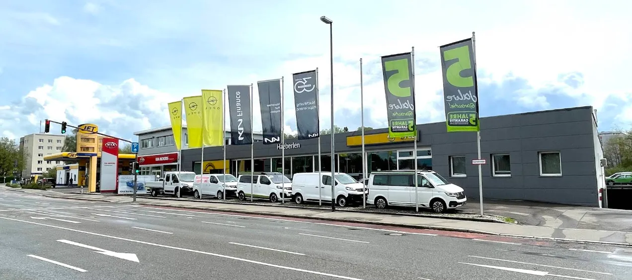 Mehrere Transporter und Autos stehen vor einem modernen Autohaus mit Stellantis- und Opel-Fahnen an einer Hauptstraße.