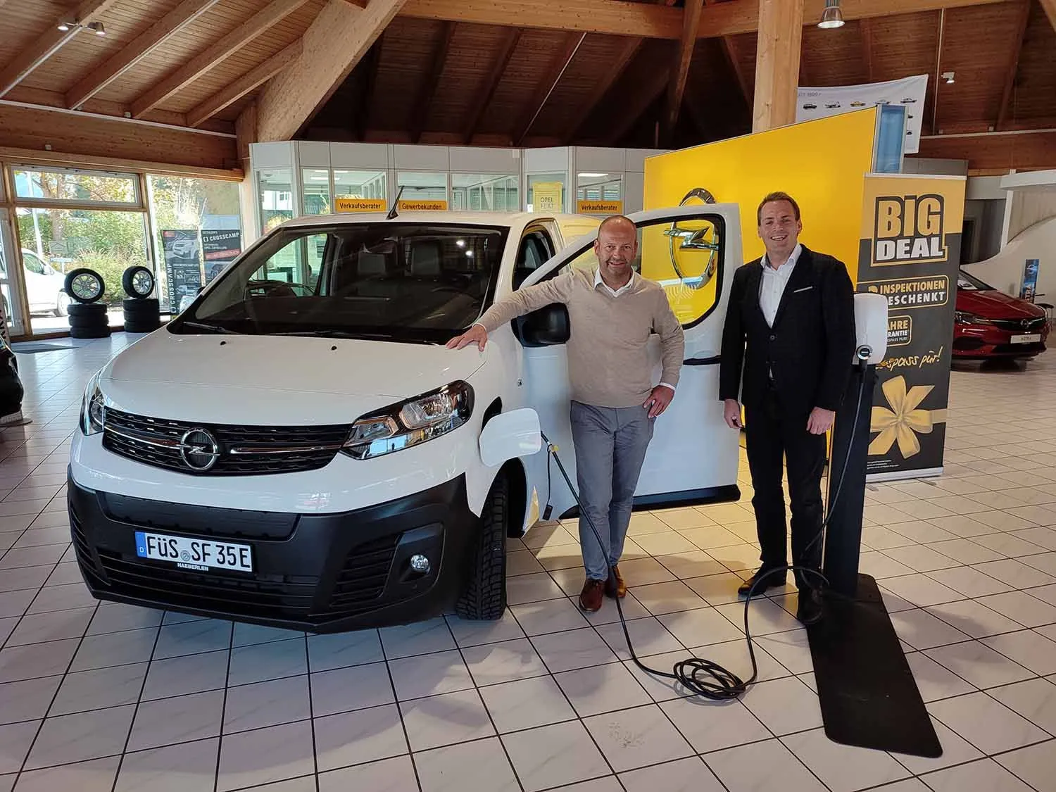 Zwei Männer präsentieren einen weißen Opel-Transporter an einer Ladestation im Autohaus, umgeben von Werbebannern.