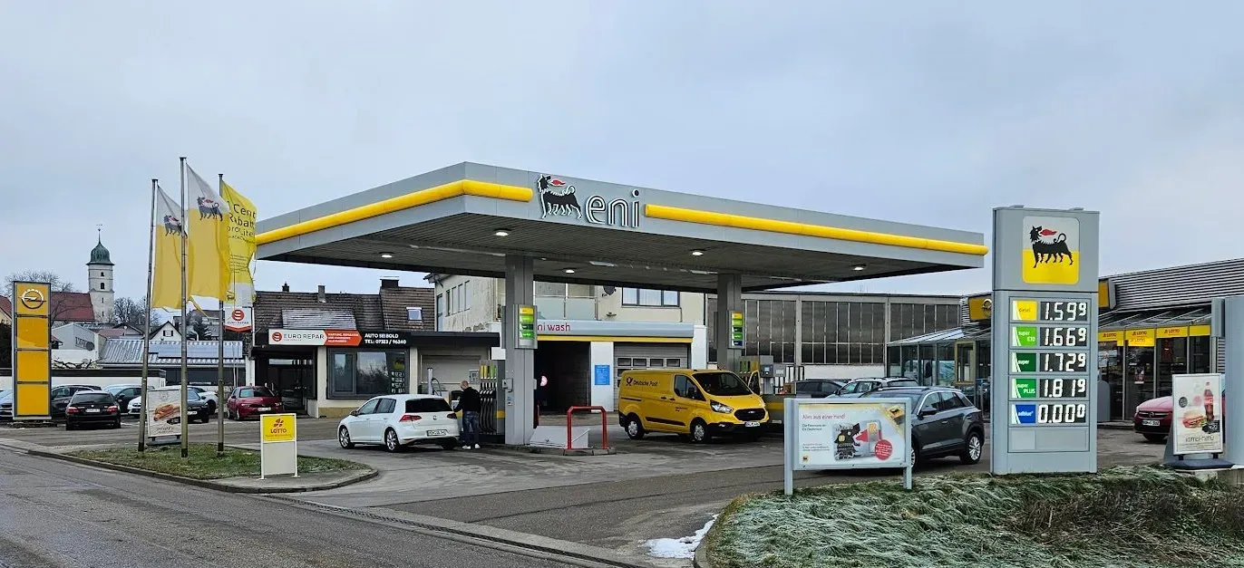 Eni-Tankstelle mit mehreren Autos, Zapfsäulen und Preisanzeige. Im Hintergrund sind Wohnhäuser und ein Kirchturm zu sehen.