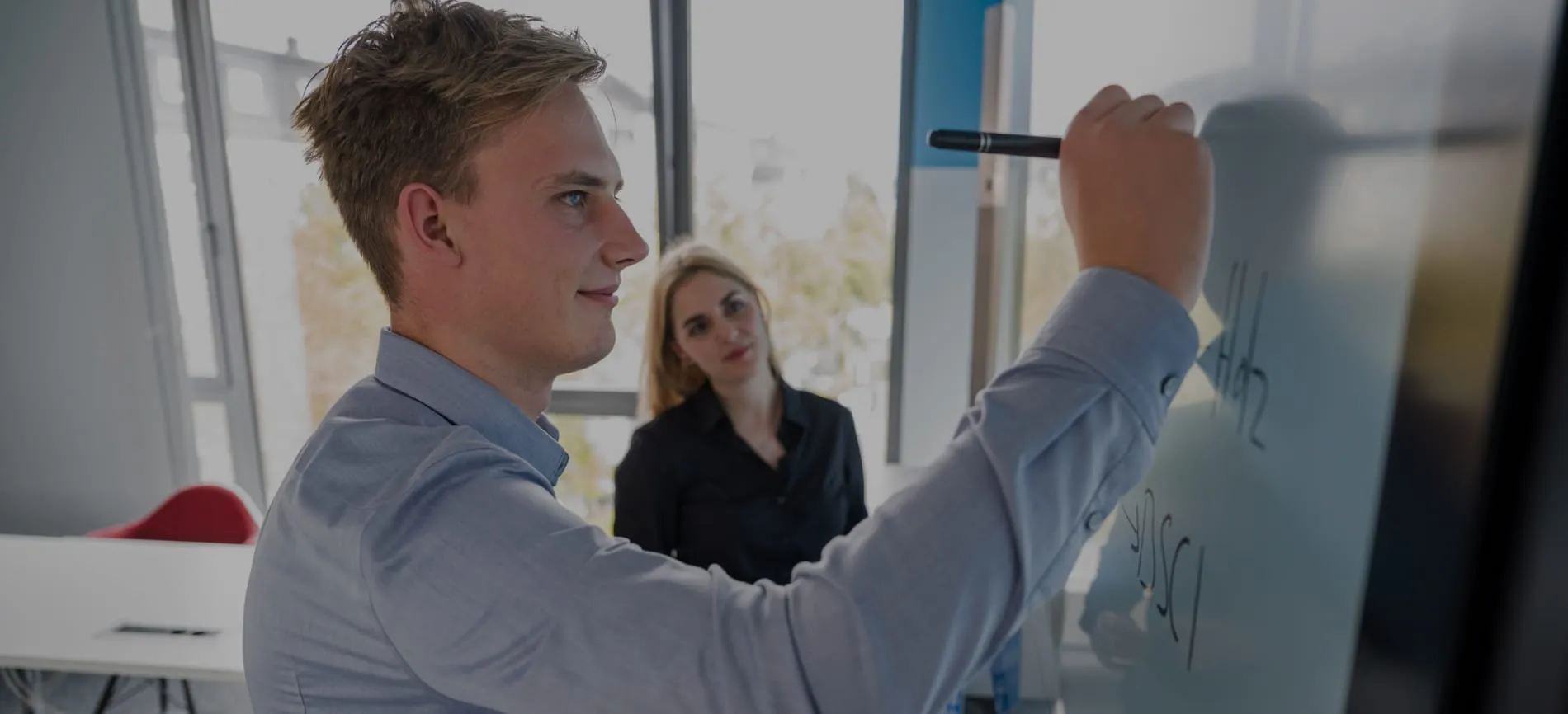 Ein junger Mann schreibt an ein Whiteboard, während eine Frau aufmerksam zuschaut, in einem modernen Büro.
