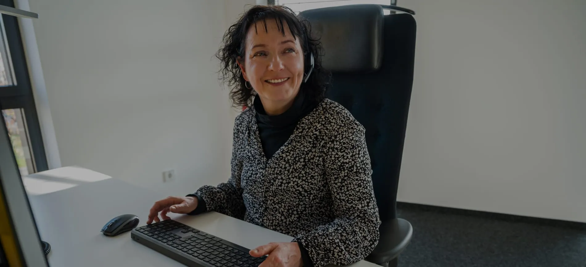 Eine lächelnde Frau mit Headset sitzt an einem Schreibtisch und tippt auf einer Tastatur in einem Büro.