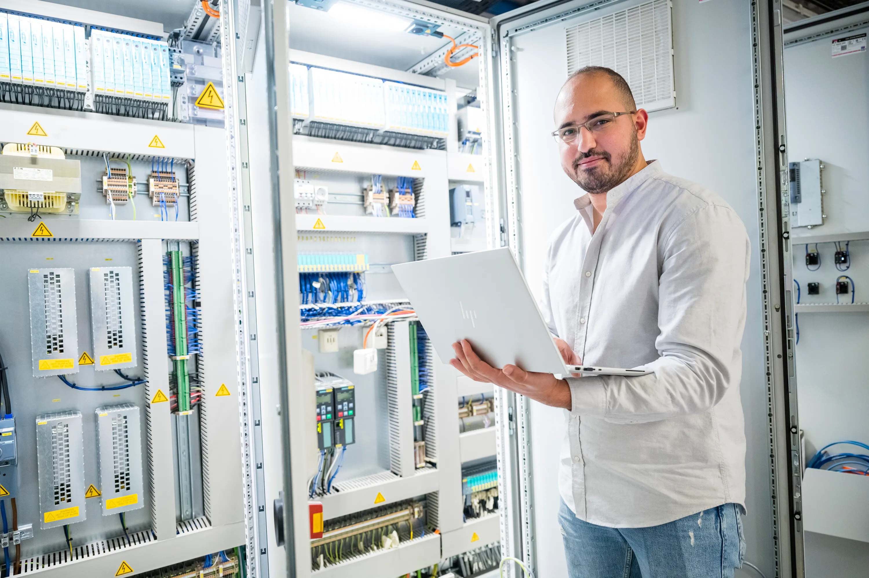 Ein lächelnder Mann in einem Serverraum hält einen Laptop in der Hand.