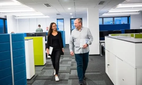 Zwei Kollegen gehen lächelnd durch ein modernes Büro bei Storch-Ciret Business Services GmbH und tragen Unterlagen.