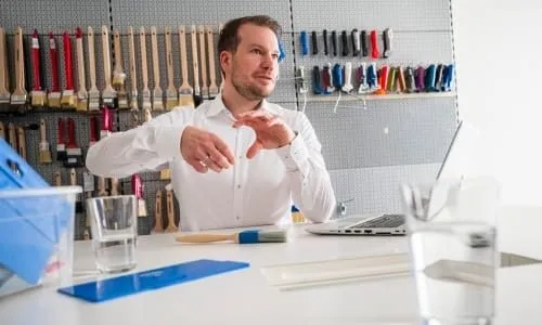 Ein Mann in einem weißen Hemd gestikuliert während eines Gesprächs in einem Büro mit Pinseln und Materialien an der Wand.