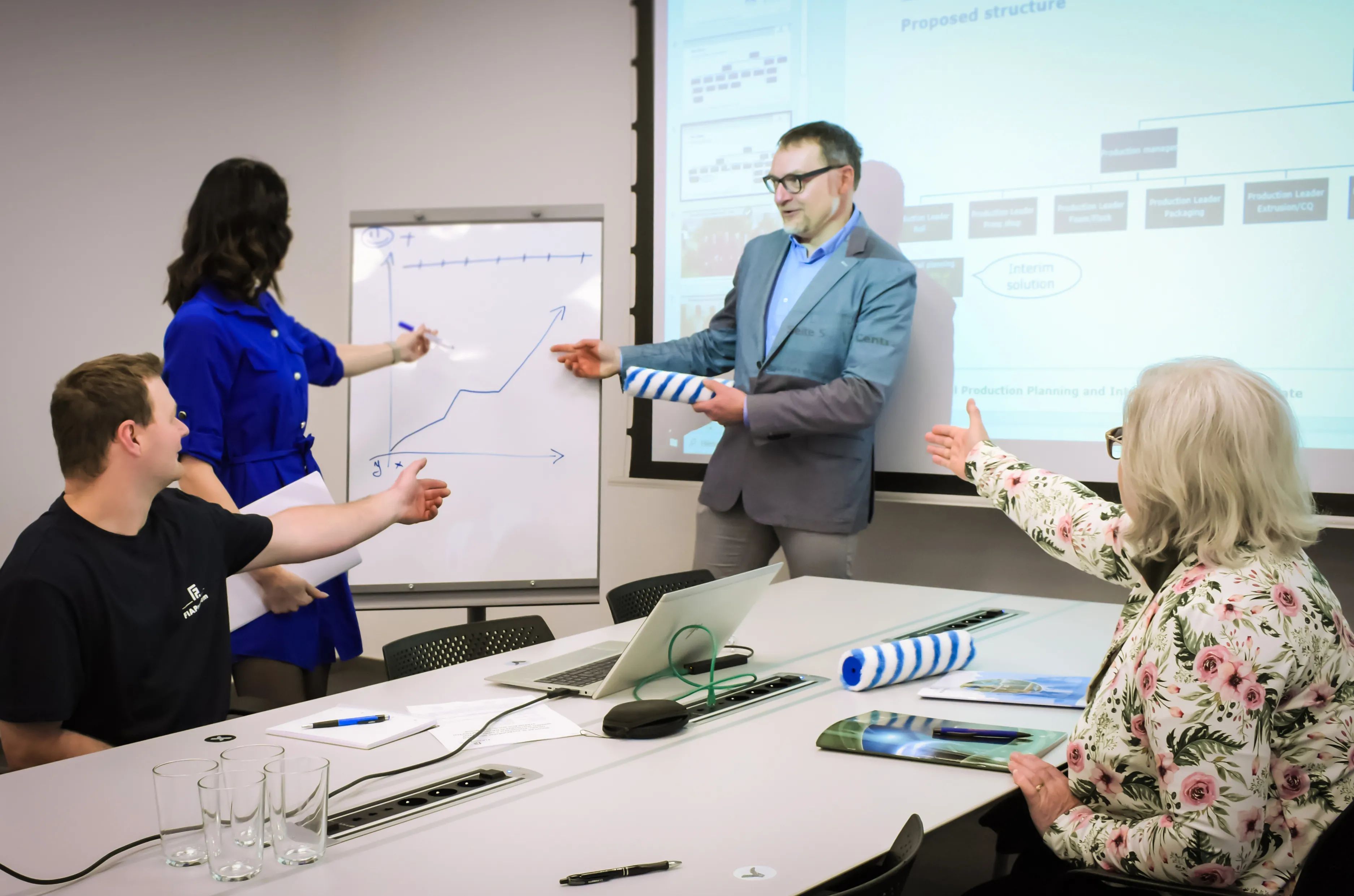 Ein Mann im weißen Hemd sitzt an einem Schreibtisch und gestikuliert, mit einem Laptop vor sich und Malerpinseln im Hintergrund.