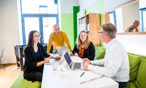 Vier Kollegen in einem modernen Büro besprechen ein Projekt an einem Tisch mit Laptops.