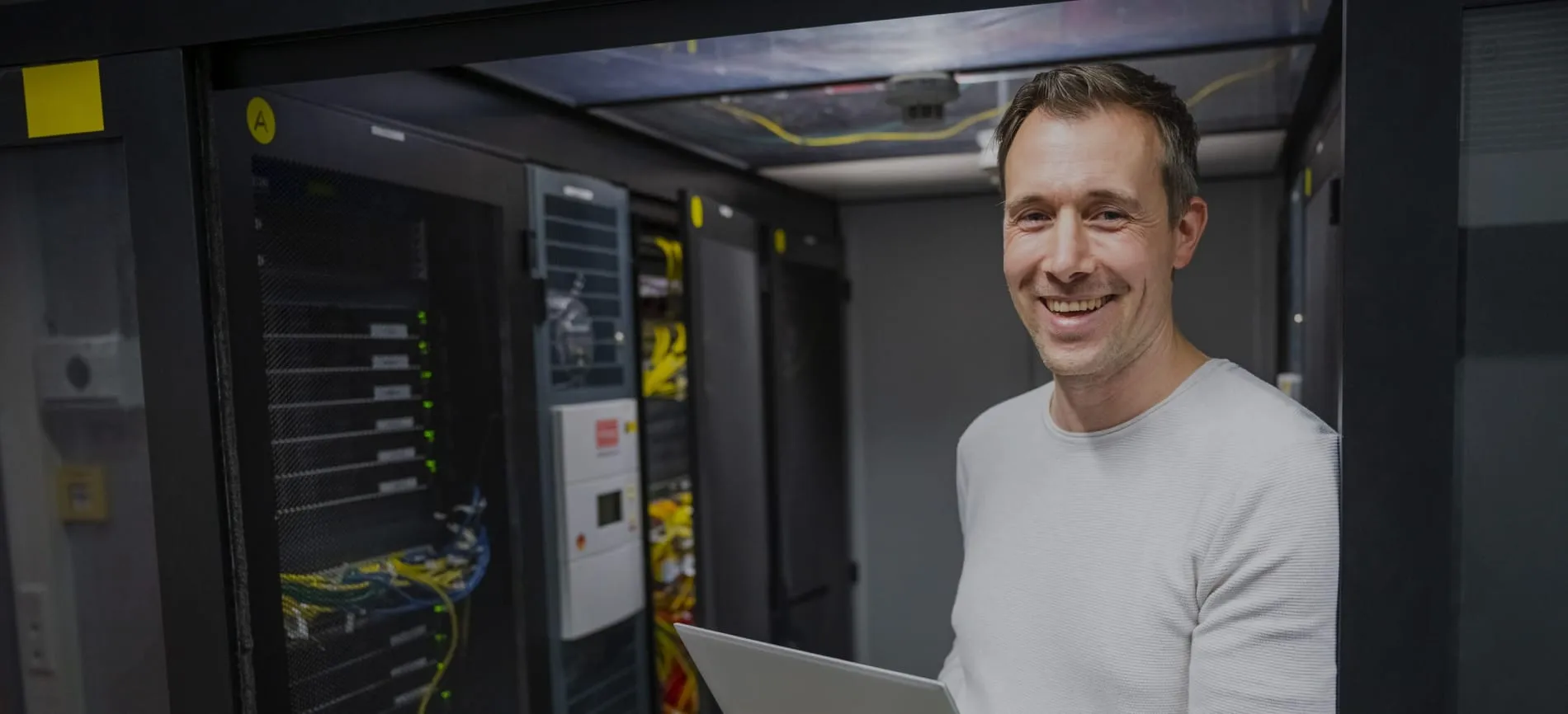 Ein lächelnder Mann steht mit einem Laptop in einem Serverraum von Storch-Ciret Business Services GmbH.