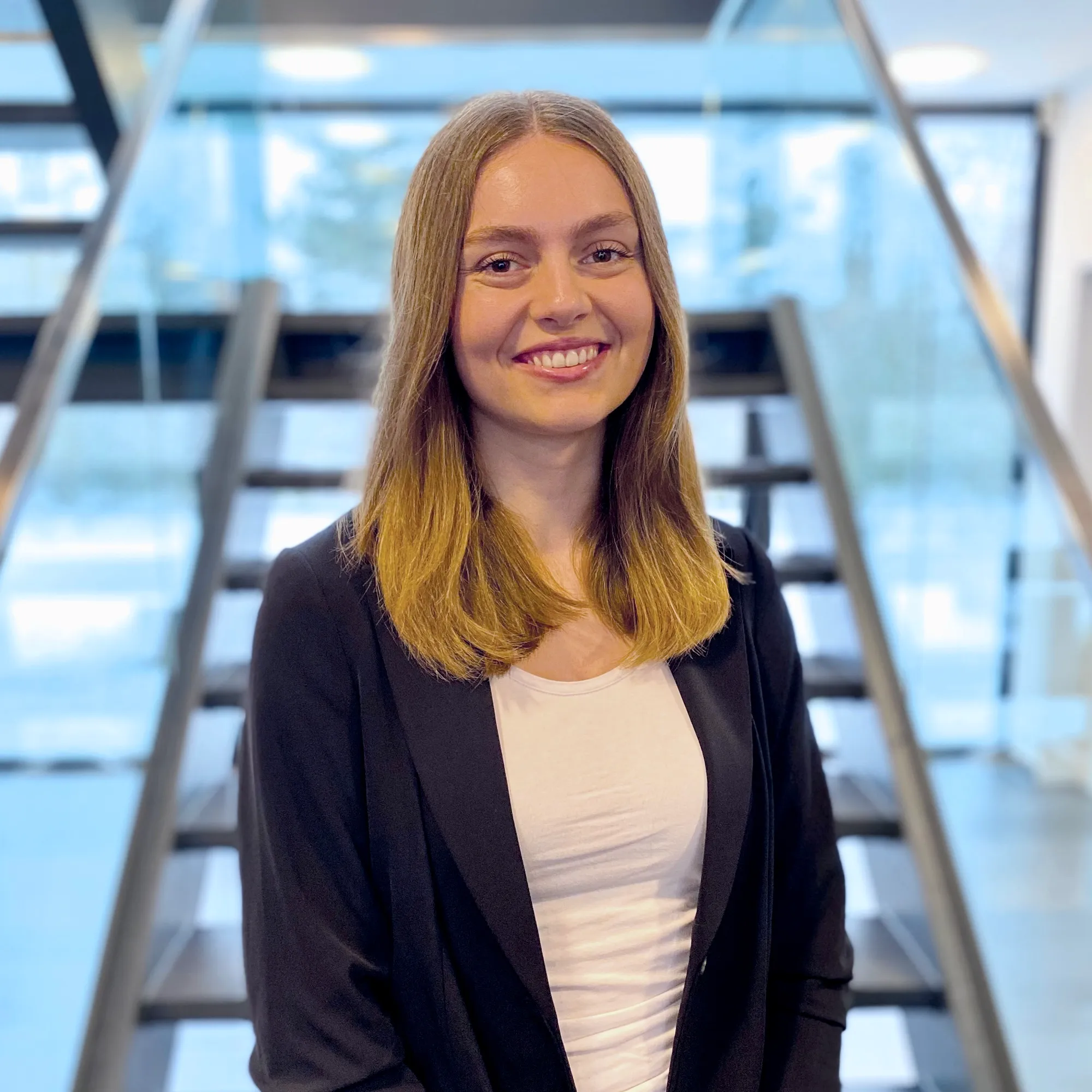 Junge Frau im Business-Outfit lächelt freundlich und steht vor einer modernen Treppe im Büro.