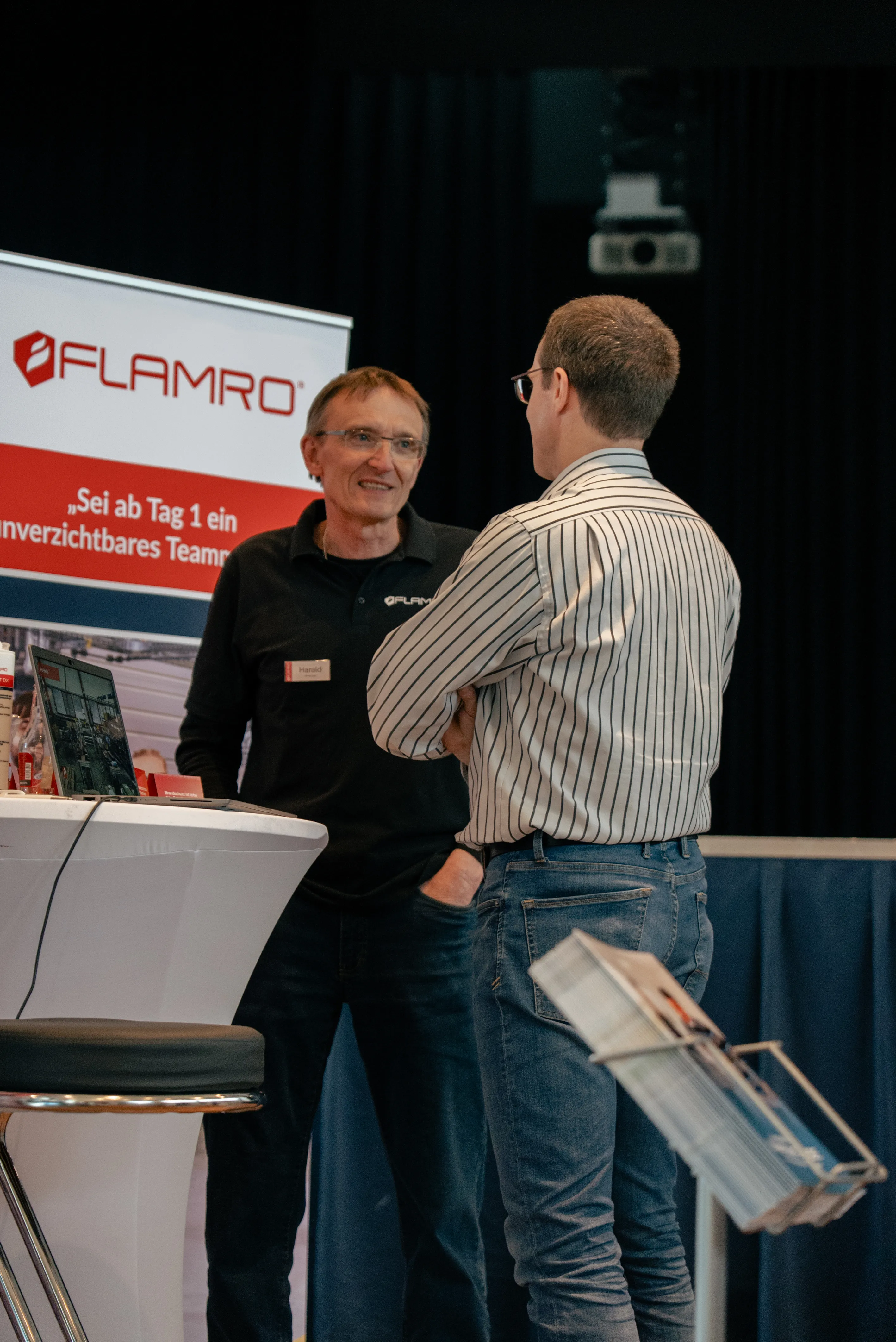 Ein Mitarbeiter von svt Holding GmbH spricht mit einem Interessenten an einem Messestand mit FLAMRO-Branding.