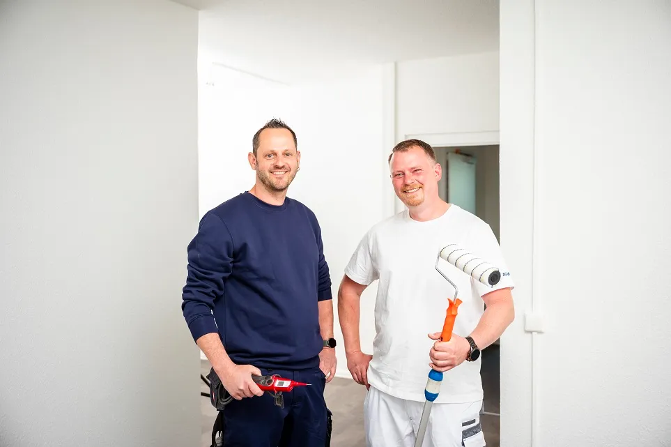 Zwei Handwerker bei TAG Immobilien AG lächeln in die Kamera, einer hält eine Farbrolle, der andere ein Werkzeug.