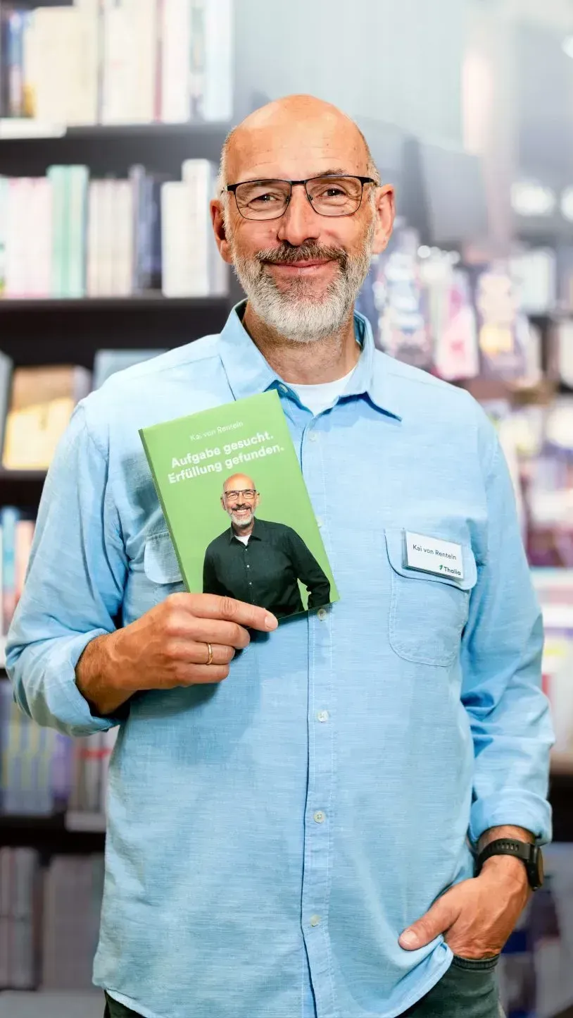 Ein freundlicher Mann mit Brille hält ein Buch in der Hand, das den Titel "Aufgabe gesucht. Erfüllung gefunden." trägt. Er steht vor einem Bücherregal und trägt ein Namensschild von Thalia.