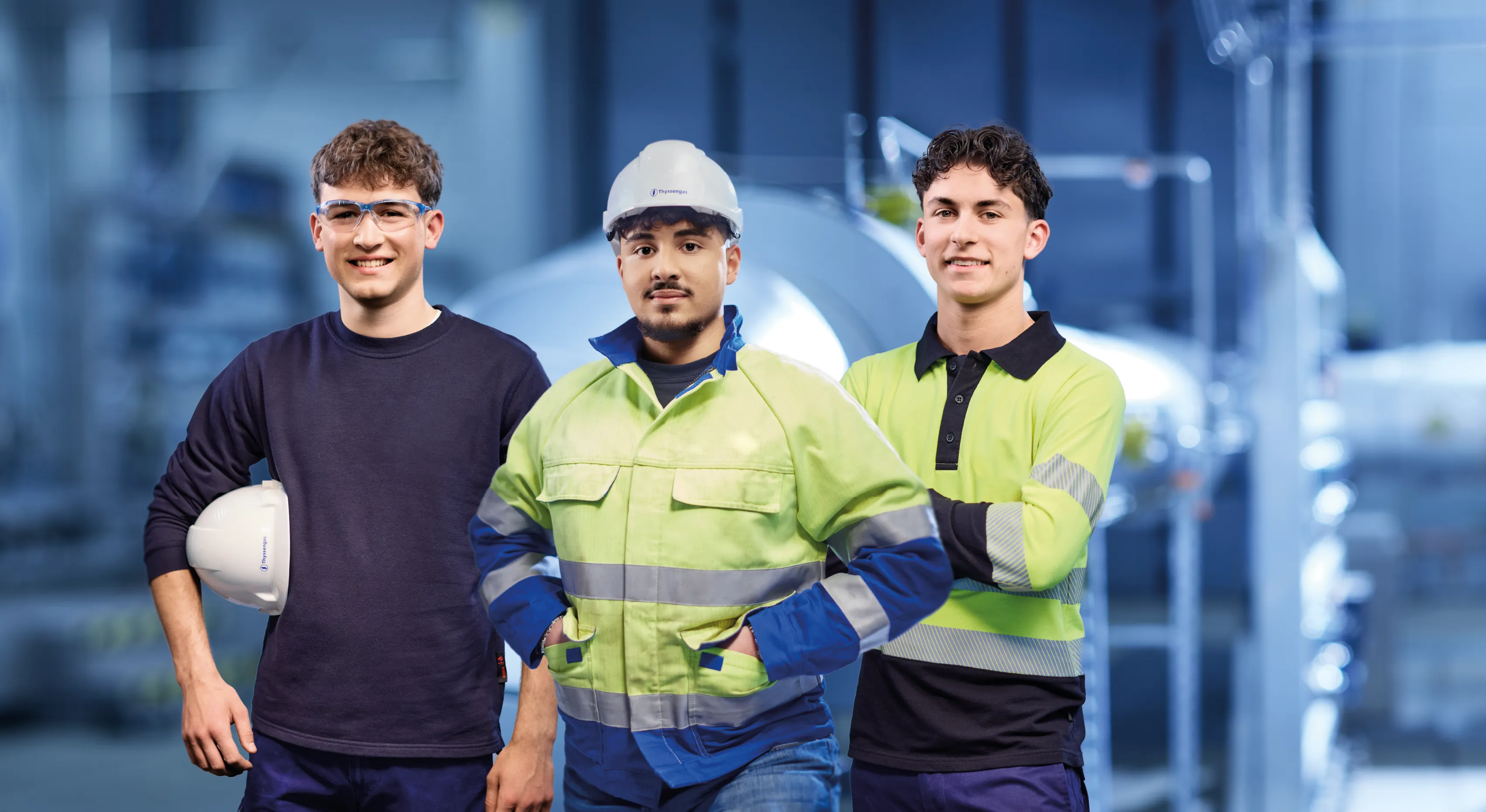 Drei junge Thyssengas-Mitarbeiter in Arbeitskleidung stehen selbstbewusst in einer Energieanlage und lächeln Dich an.