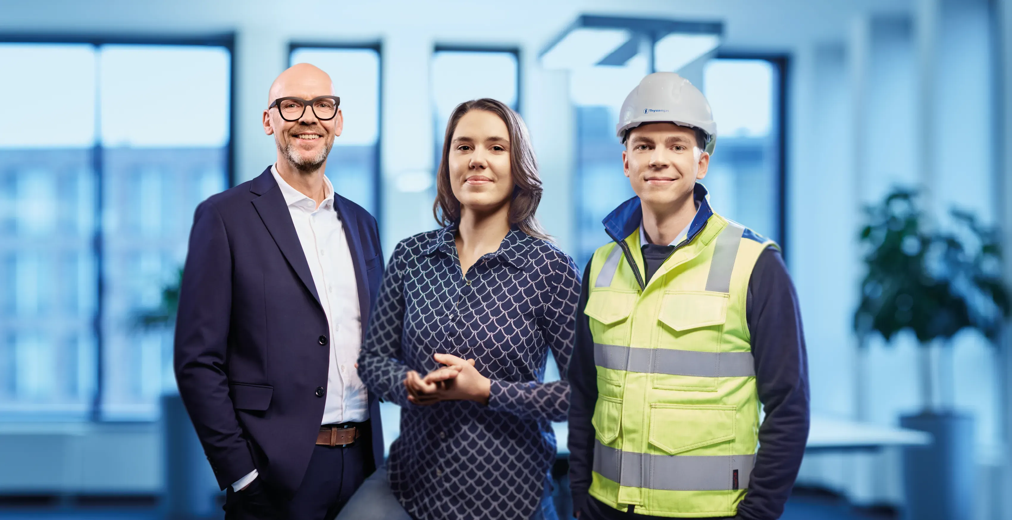 Drei Thyssengas-Mitarbeiter aus Büro und Technik stehen gemeinsam in einem modernen, hellen Büro und lächeln dich an.