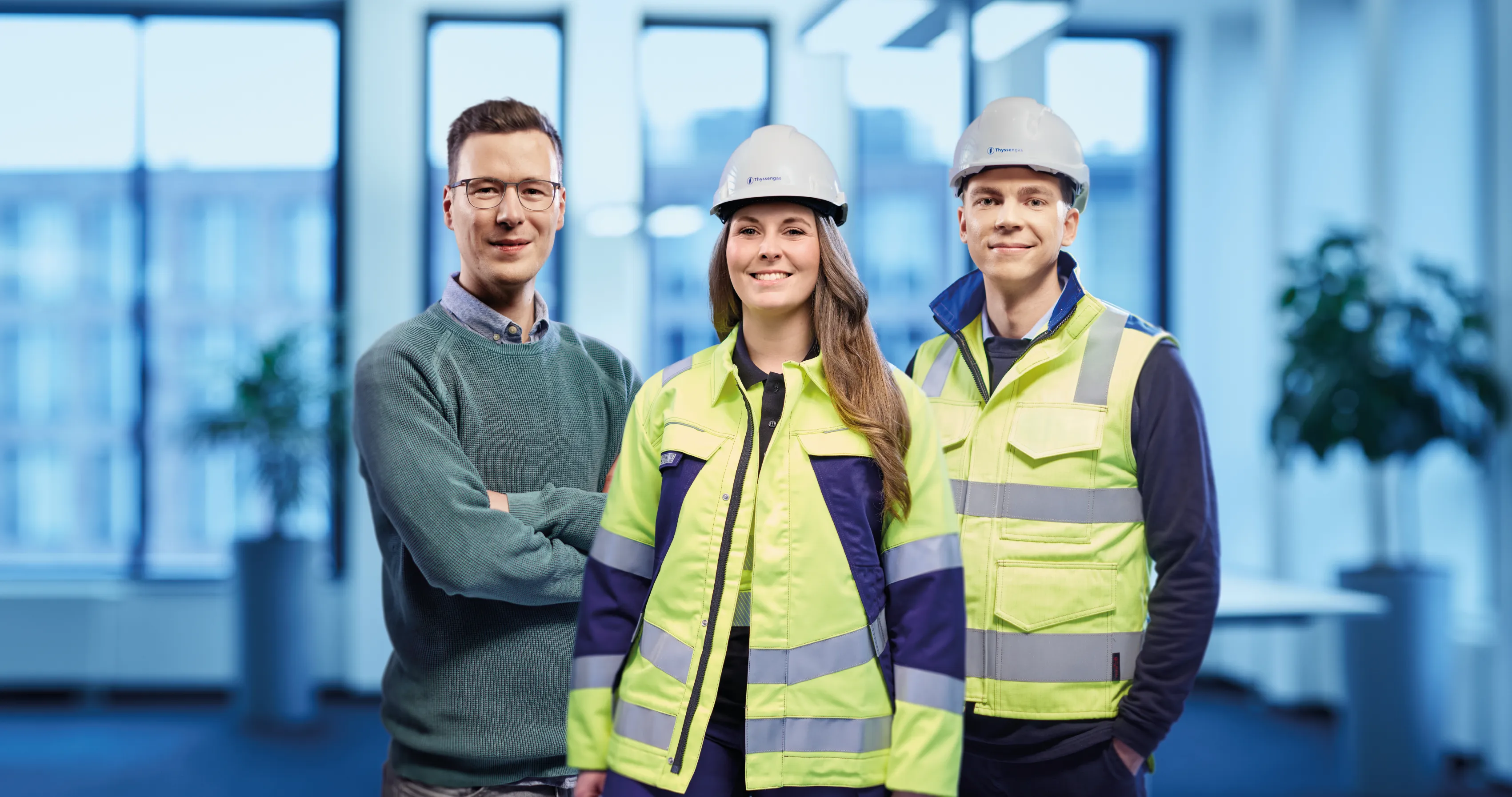 Drei junge Thyssengas-Mitarbeitende, zwei in Arbeitskleidung und Helm, einer im Pullover, stehen gemeinsam im Industrieumfeld.