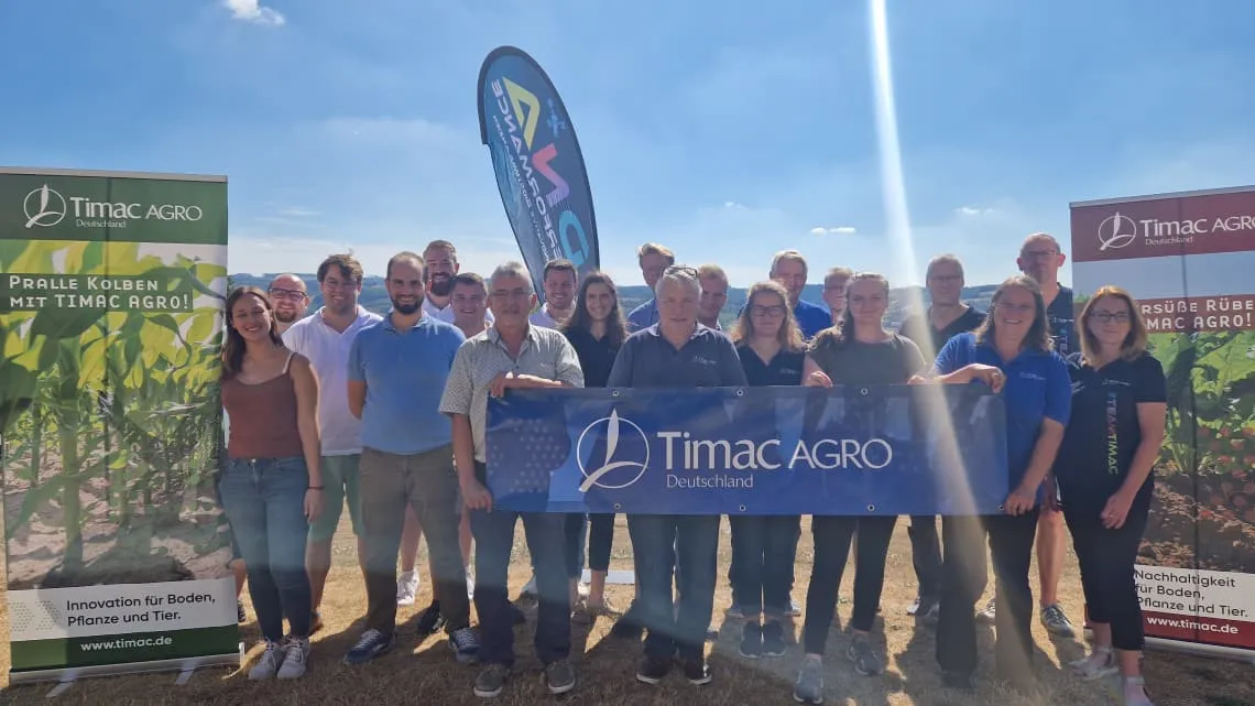 Ein Team von TIMAC AGRO Deutschland steht lächelnd im Freien und hält ein Firmenbanner bei sonnigem Wetter.