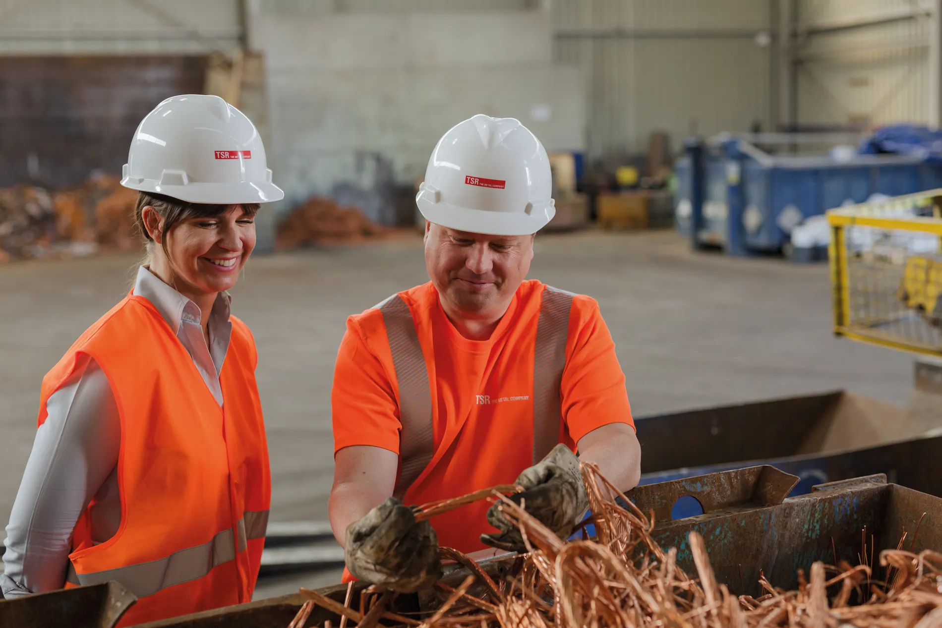 Zwei Mitarbeiter von TSR Group GmbH & Co. KG in einer Lagerhalle prüfen Kupfermaterial, tragen Schutzhelme und Sicherheitswesten.